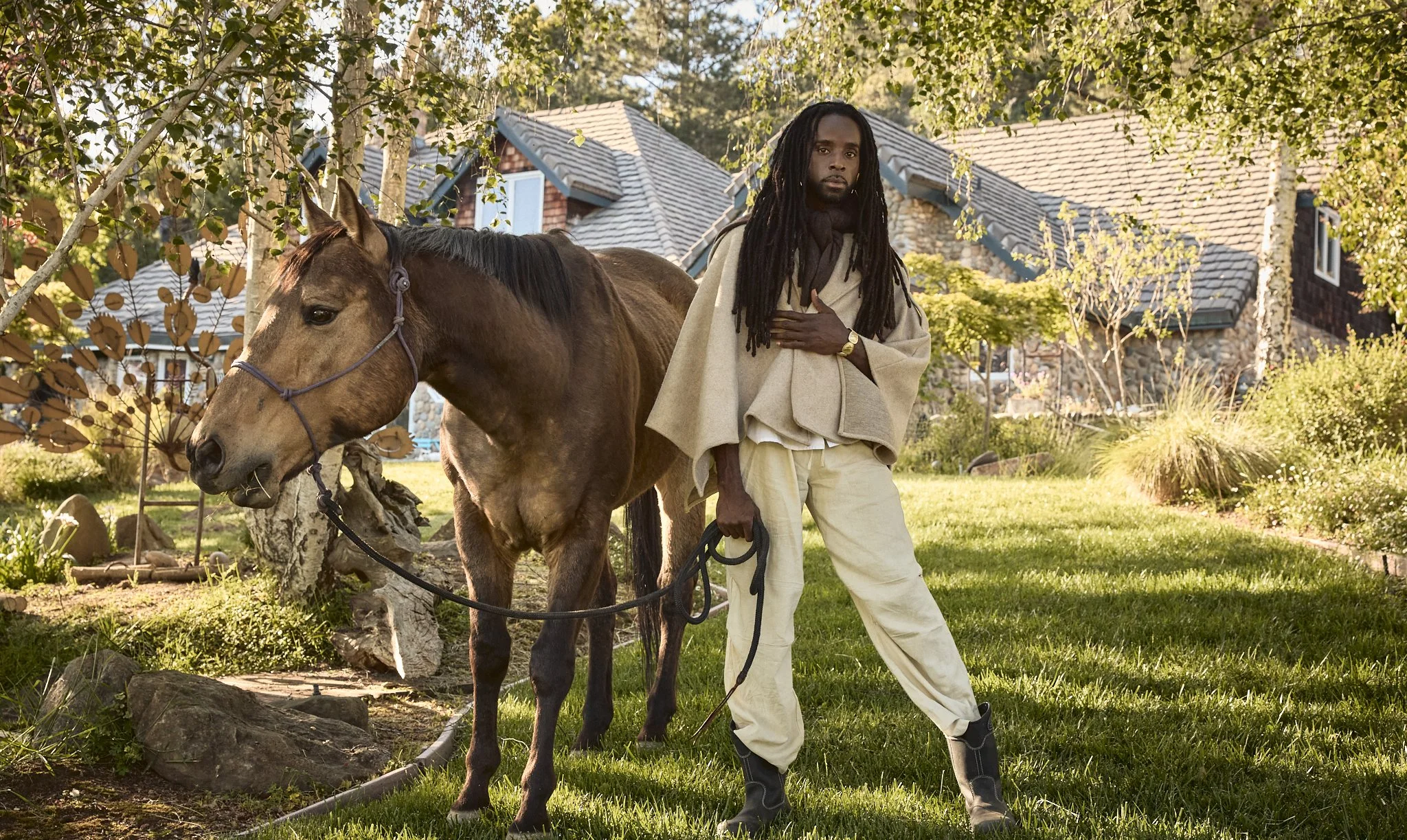 A man with long dreadlocks, wearing a beige cloak and loose pants, stands next to a brown horse on a grassy lawn in a residential area with stone houses and trees.