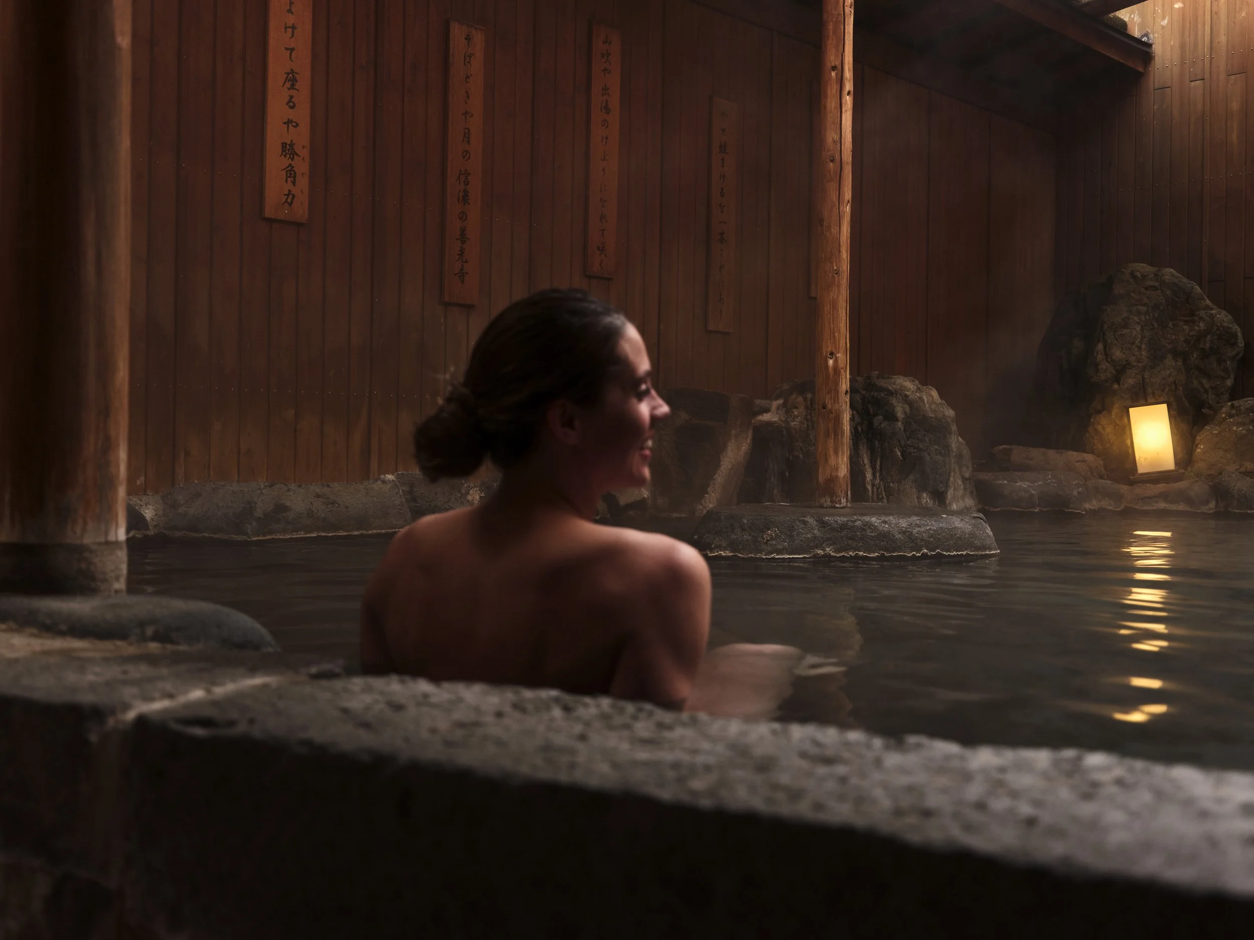 A woman relaxing in a wooden hot spring bath at dusk, with warm lighting and Japanese wooden plaques on the wall.