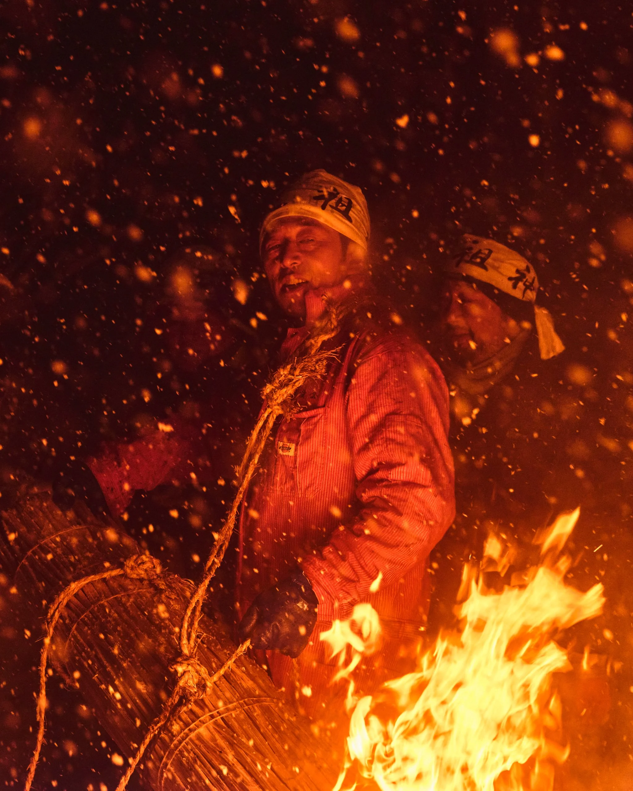 Person wearing a headband and jacket, standing amidst flames and sparks, holding a rope tied around logs during a fire-related activity at night.