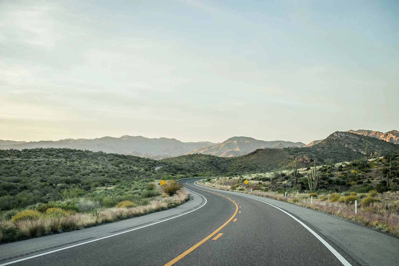 Winding asphalt road through desert landscape with green shrubs, cacti, and mountains in the background under a partly cloudy sky.