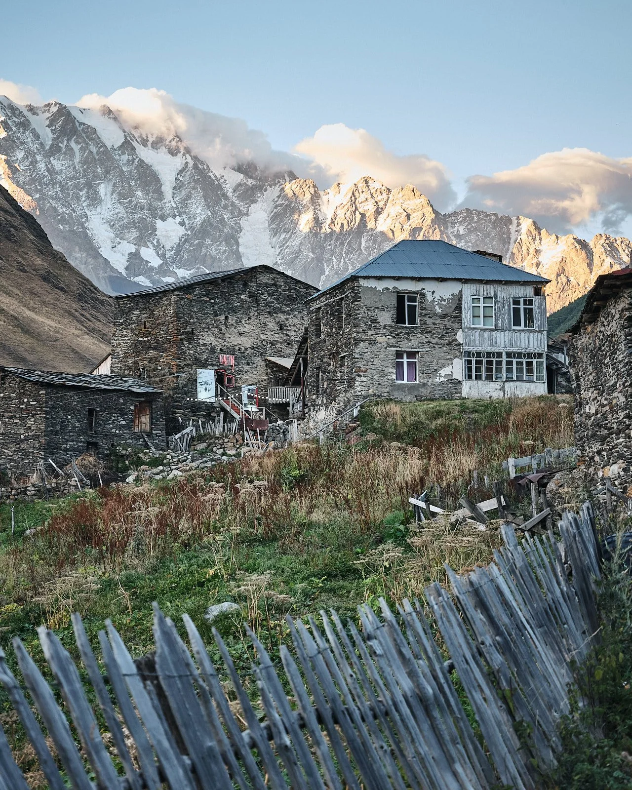 A rustic mountain village with stone houses, surrounded by tall, snow-capped mountains and a partly cloudy sky.