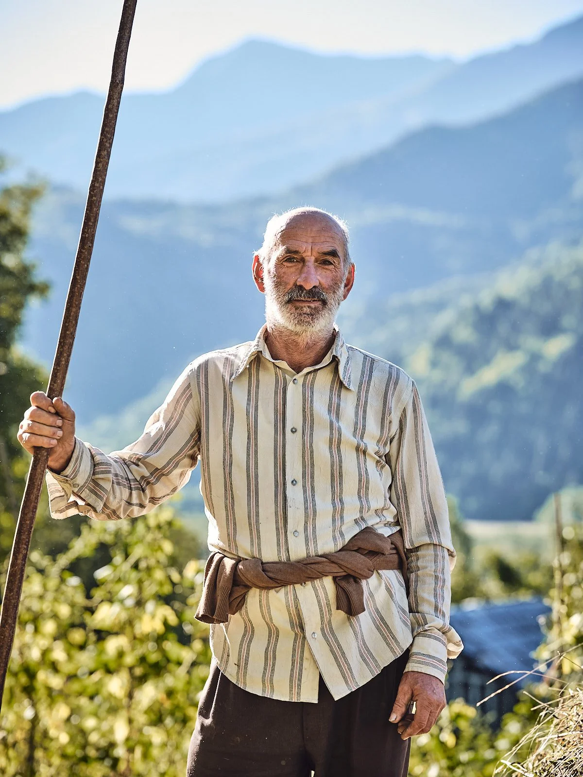 An elderly man standing outdoors in a rural area with mountainous landscape in the background, holding a stick, dressed in a striped shirt and dark pants with a brown sweater tied around his waist.