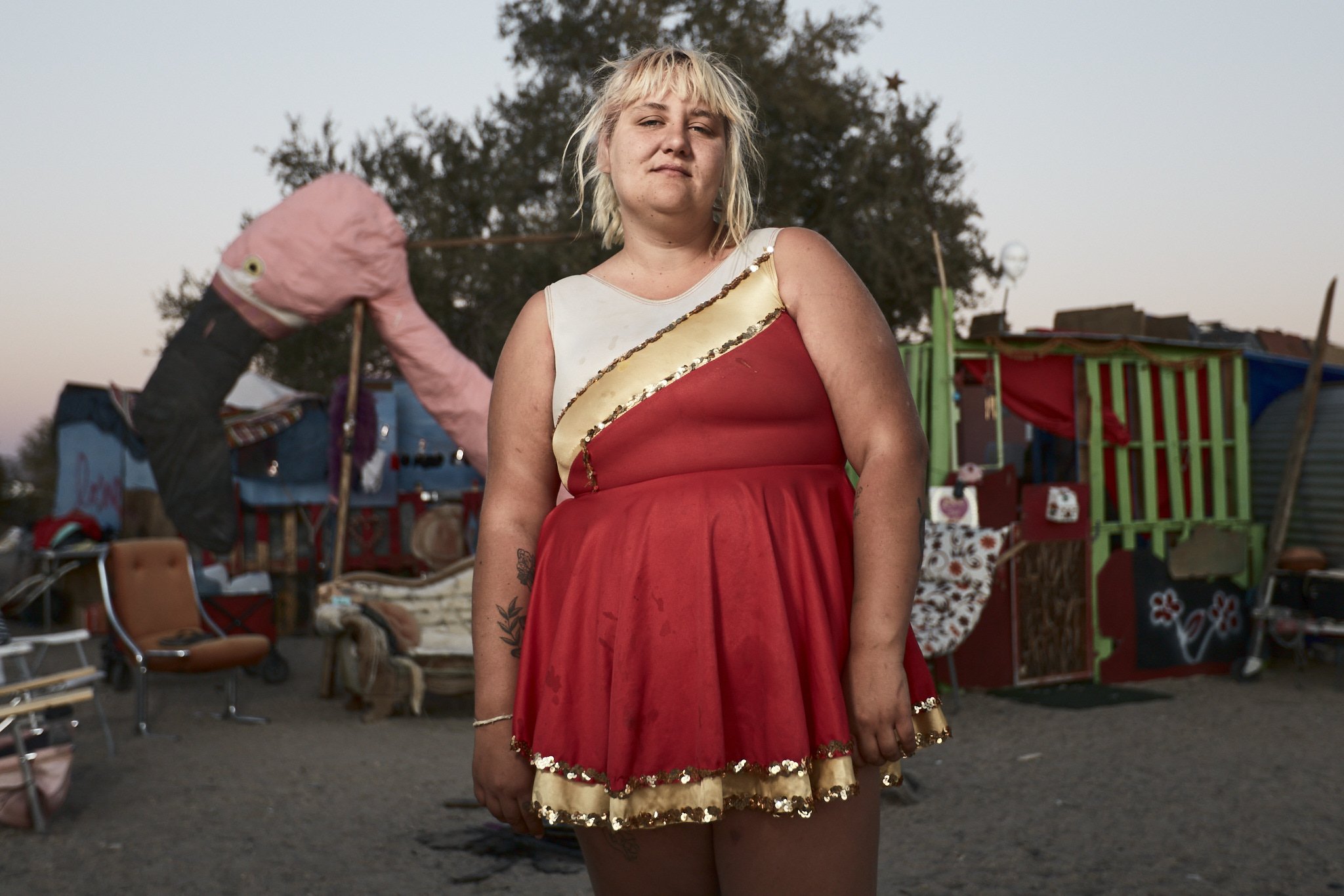 A woman with blonde hair wearing a red dress with a gold stripe stands in front of a colorful, makeshift outdoor area with furniture and a large pink flamingo decoration.