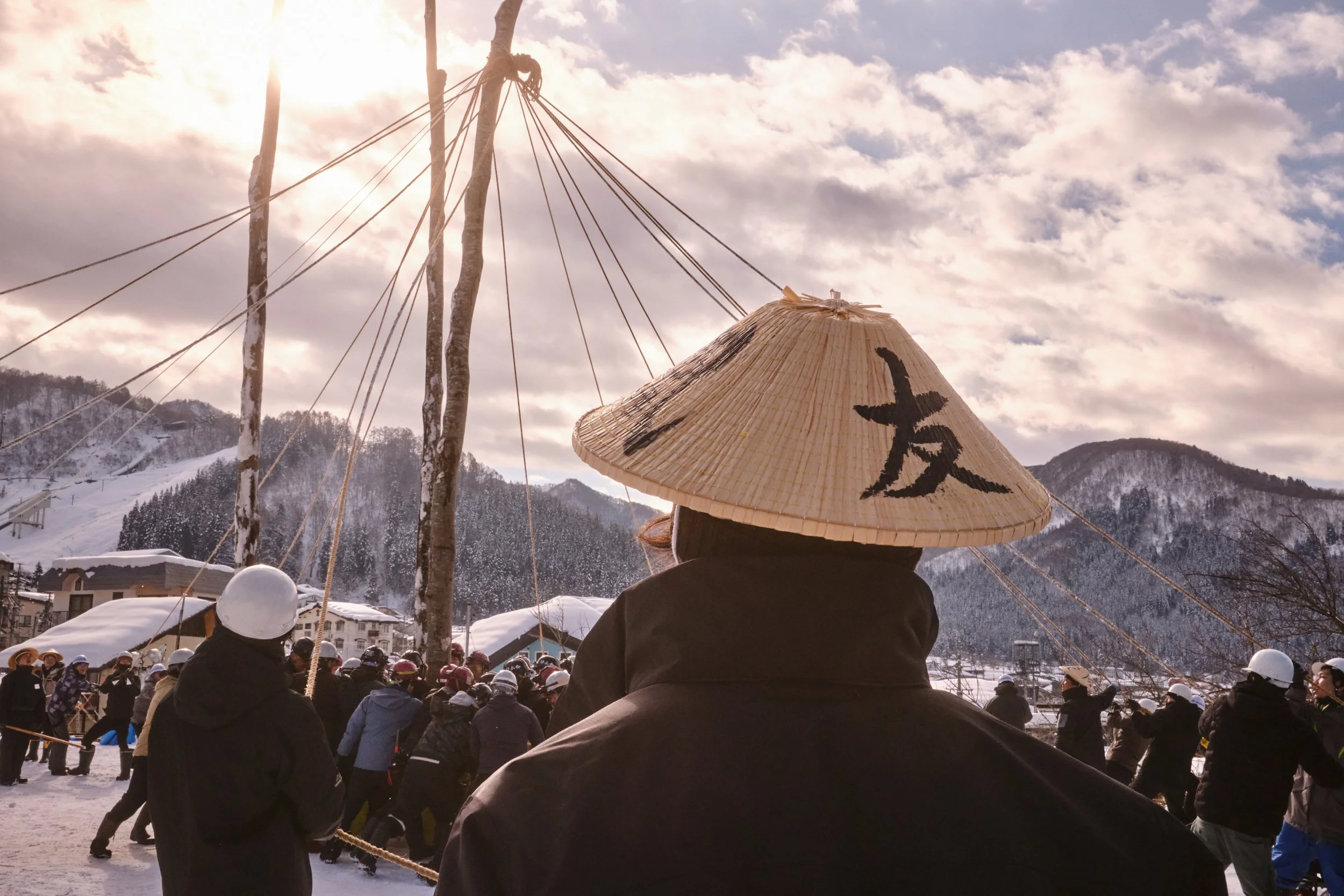 A person wearing a traditional Asian conical hat with Chinese characters, with a crowd in the background, snow-covered mountains, and a cloudy sky.