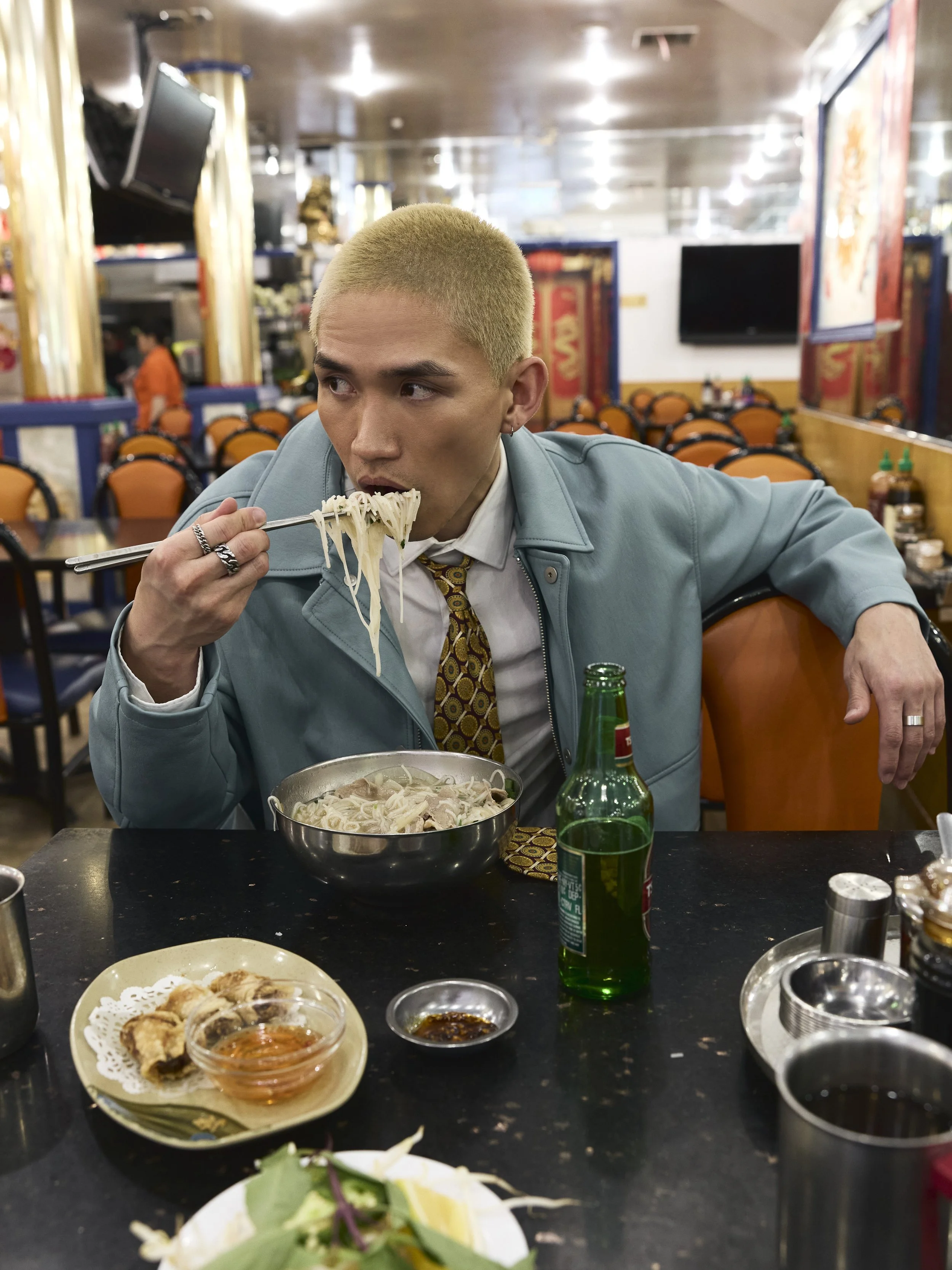A man with short blond hair wearing a blue jacket, white shirt, and patterned tie, eating noodles with chopsticks in a restaurant. There are various condiments and dishes on the table, and the restaurant has a colorful decor with orange chairs and mu