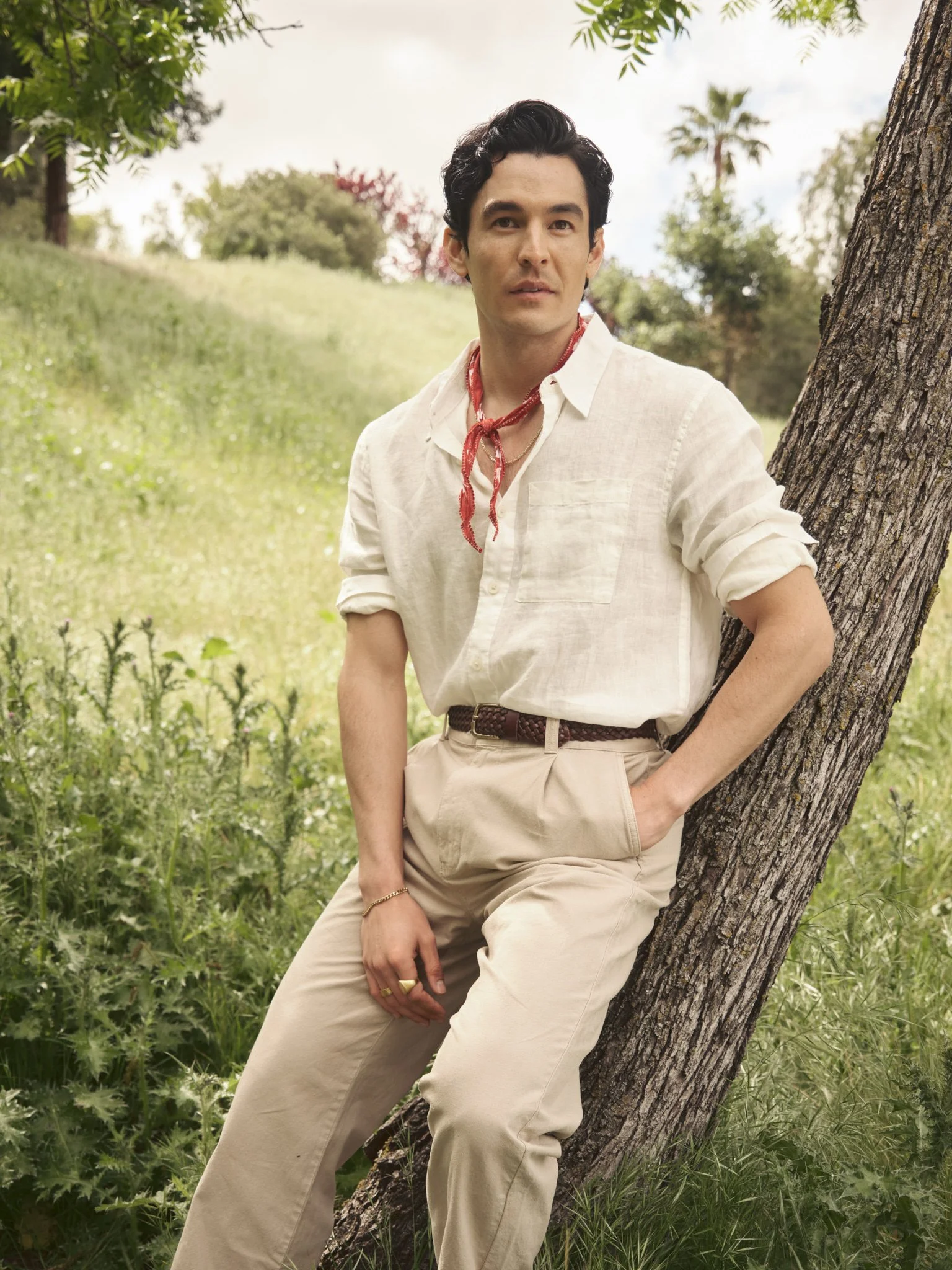 A man leaning against a tree outdoors in a grassy area with trees and a cloudy sky in the background, wearing a white shirt, beige pants, a red bandana, and jewelry.