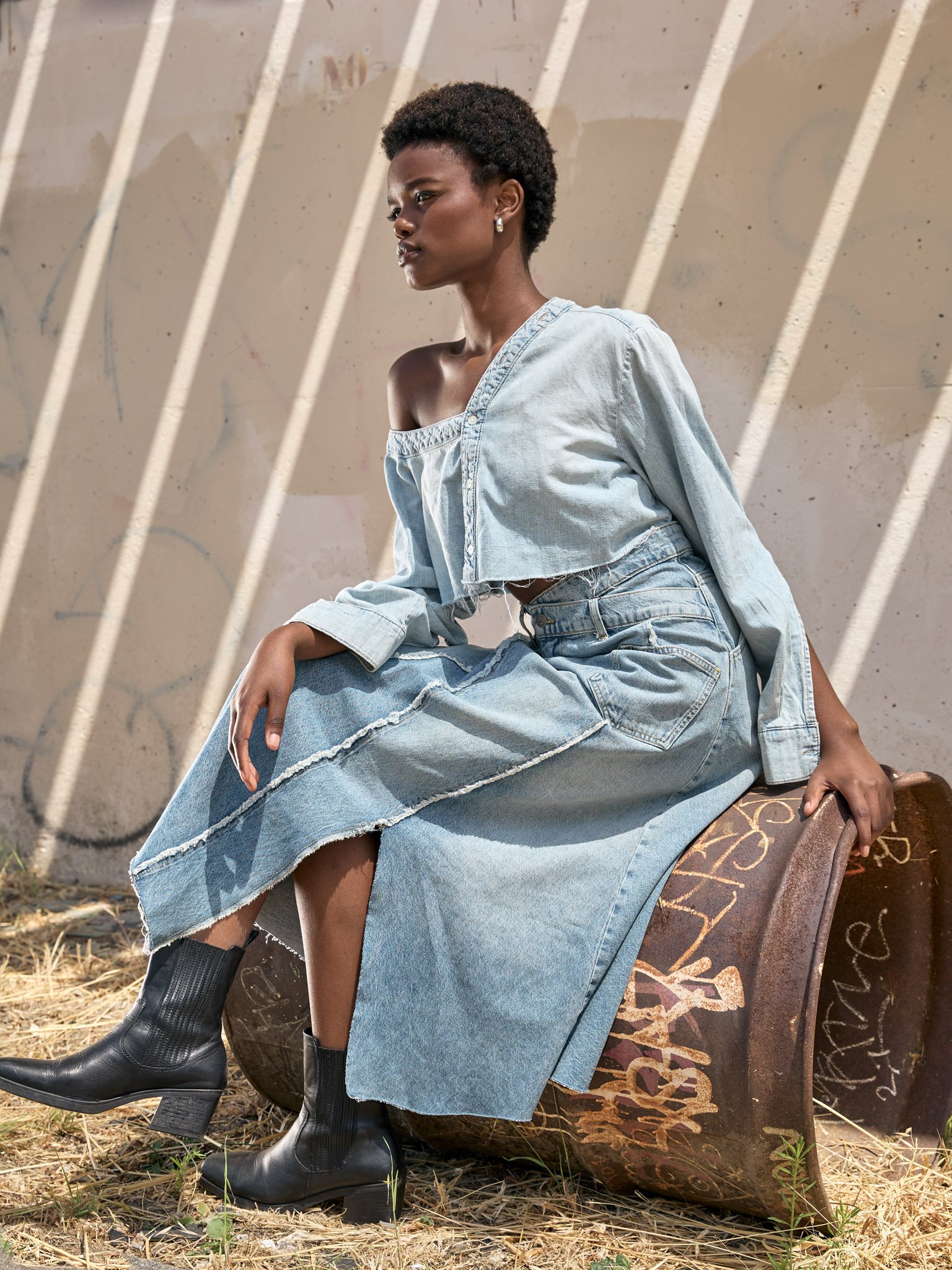 A woman with dark skin and short curly hair, wearing a light blue off-the-shoulder denim top and long denim skirt, sitting on a rusty barrel with graffiti, outdoors on dry grass, with graffiti-covered wall in background.