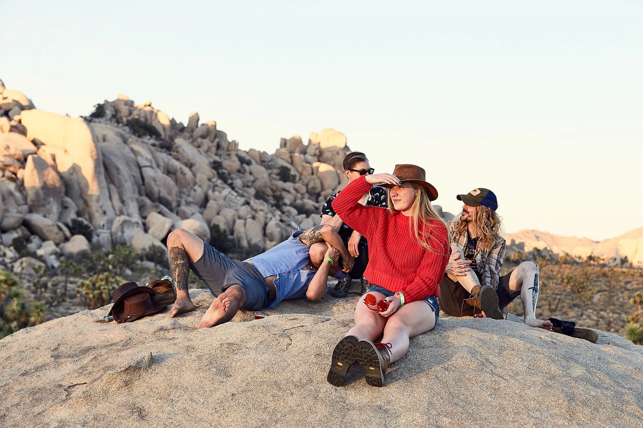 Four people sitting and relaxing on a large rock in a desert landscape with rocky mountains in the background, during sunset.