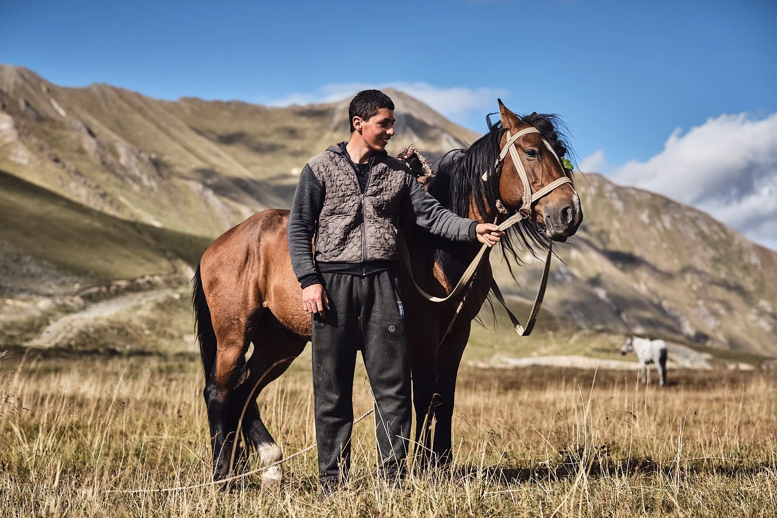 A young man standing next to a brown horse in a grassy field with mountains in the background, holding the horse's bridle in his hand.