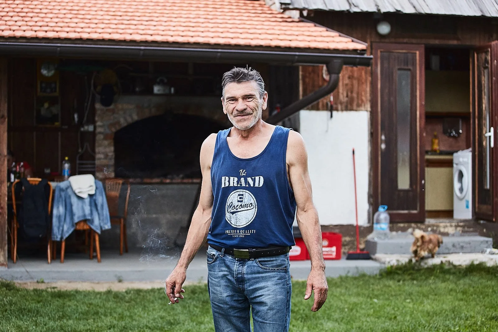 An older man with gray hair and a beard wearing a sleeveless blue tank top and jeans standing on a lawn in front of a rustic house with a patio, laundry, and a small dog in the background.