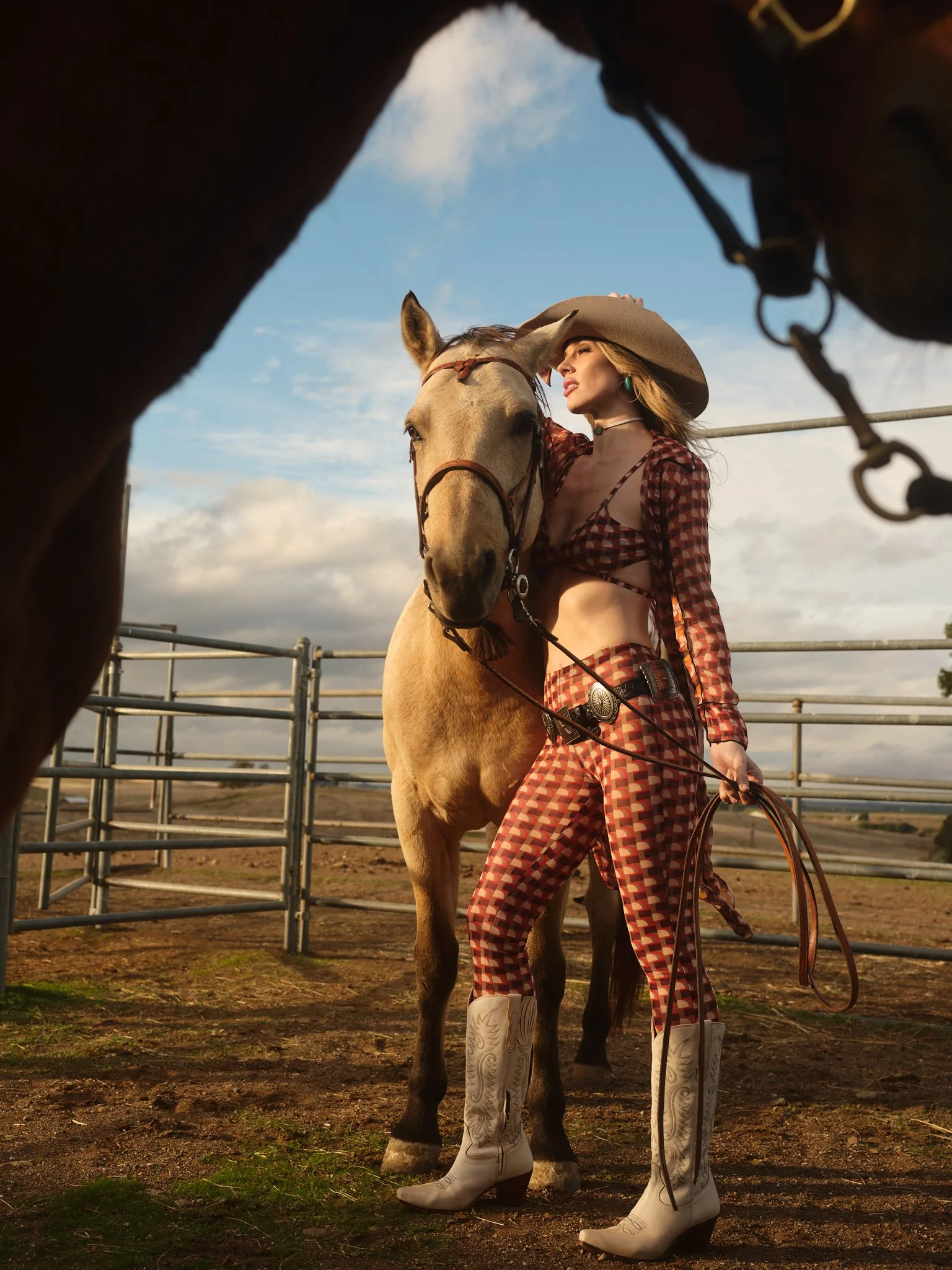 A woman in rodeo attire standing next to a light-colored horse in an enclosure during daytime, with a partly cloudy sky in the background.