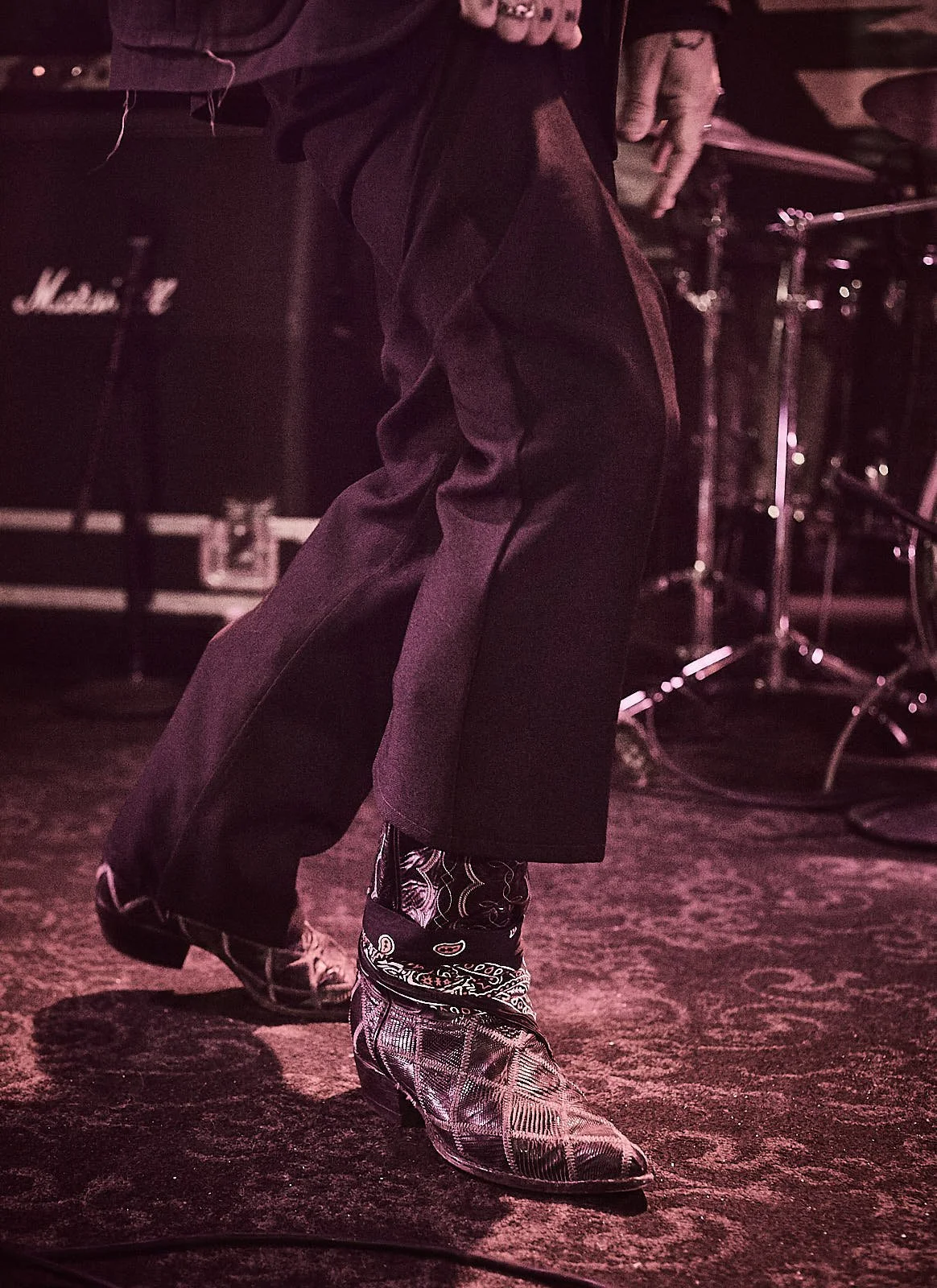 Person dancing or performing on stage, wearing shiny patterned cowboy boots, dark pants, and a dark jacket, with musical equipment like drums and amplifiers in the background.