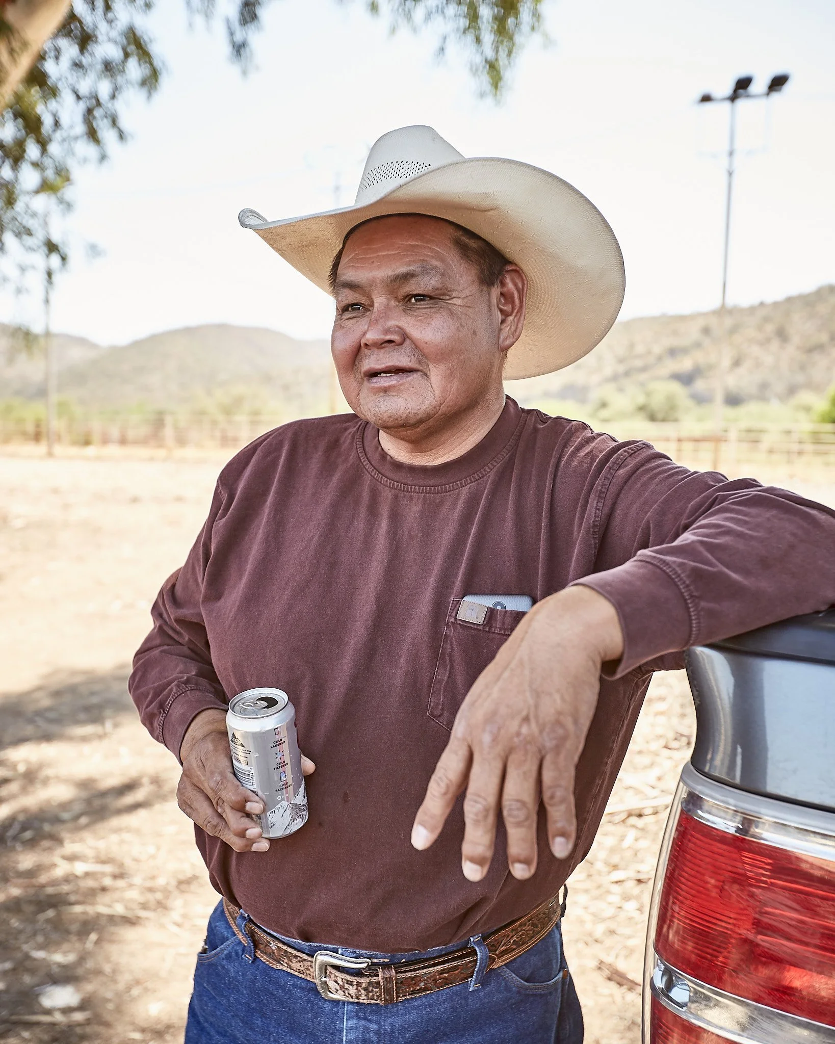 An older man wearing a cowboy hat and maroon long-sleeve shirt holding a soda can, leaning against the back of a car in an outdoor rural setting with hills in the background.