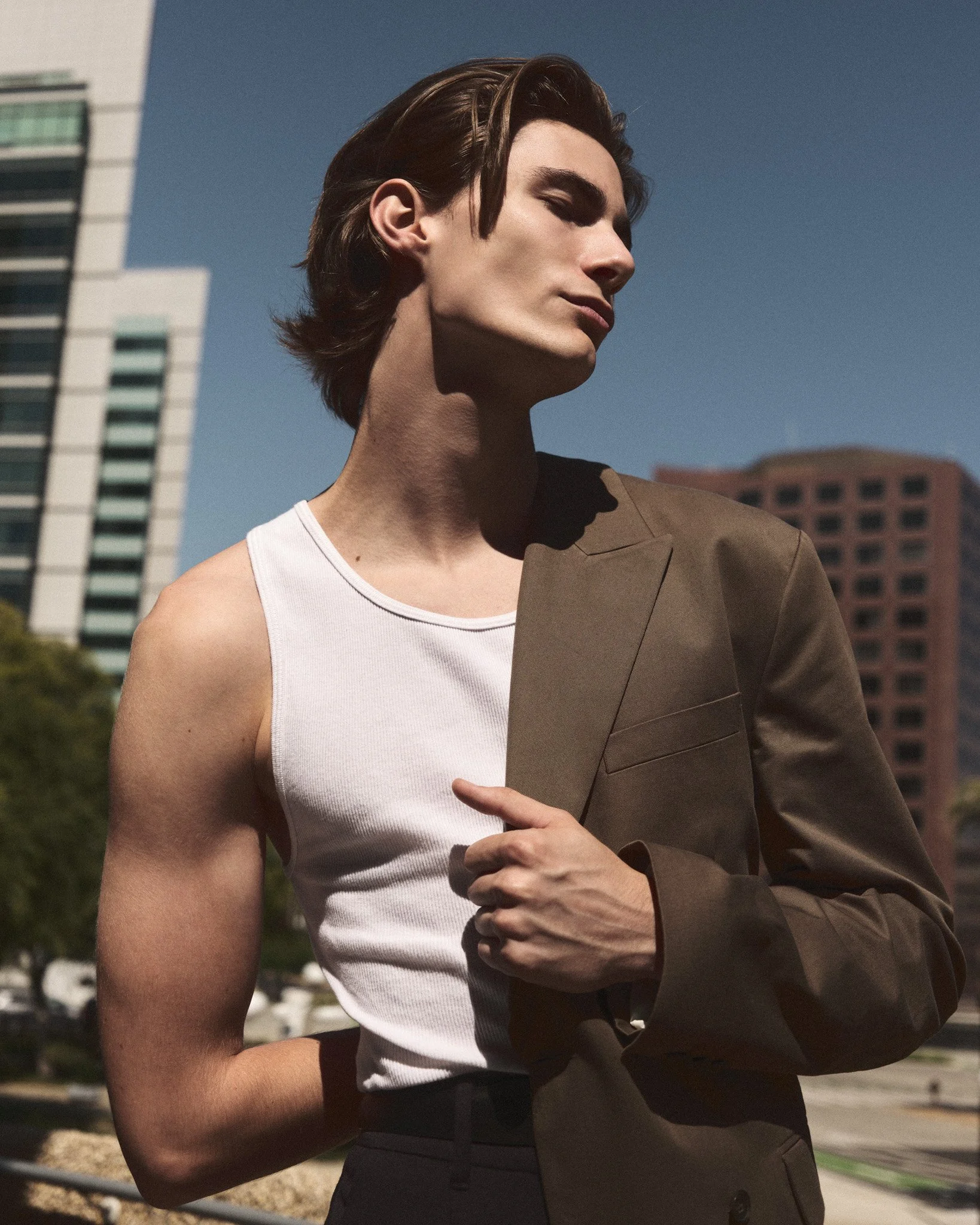 A young man with styled hair, wearing a white sleeveless shirt and a tan blazer draped over his shoulder, standing outdoors in front of modern buildings under a clear sky.