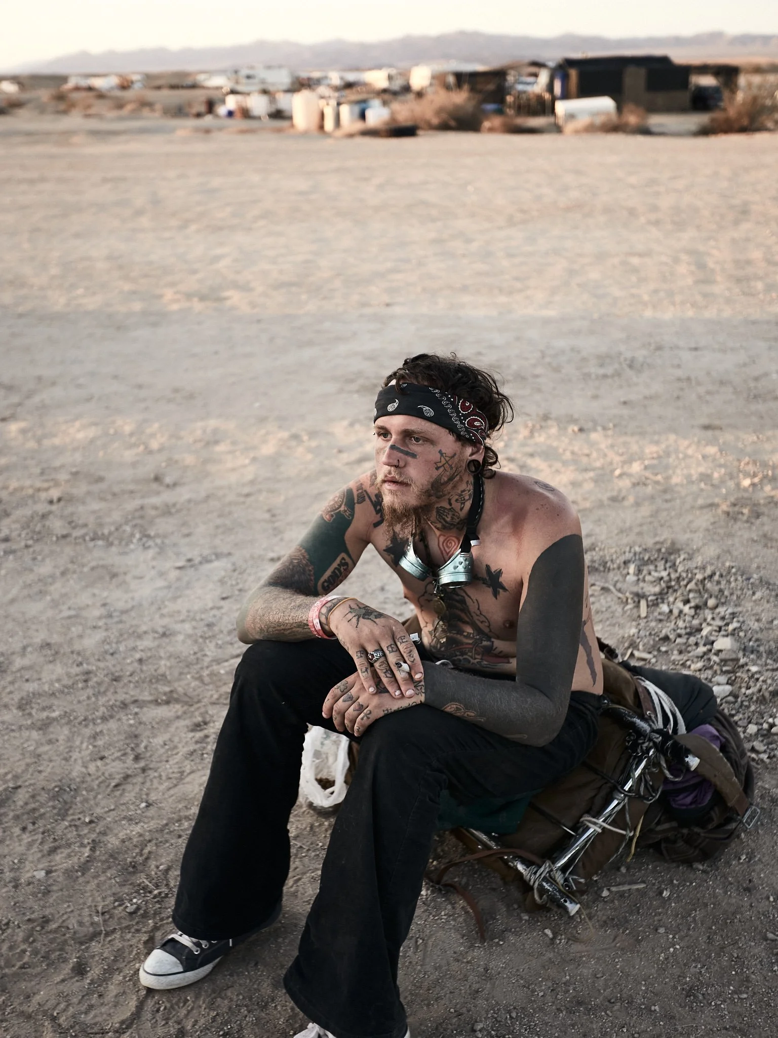 A shirtless man with numerous tattoos sitting on a dirt ground in a desert, with a backpack beside him and a scenic background of desert landscape with distant trailers or buildings.