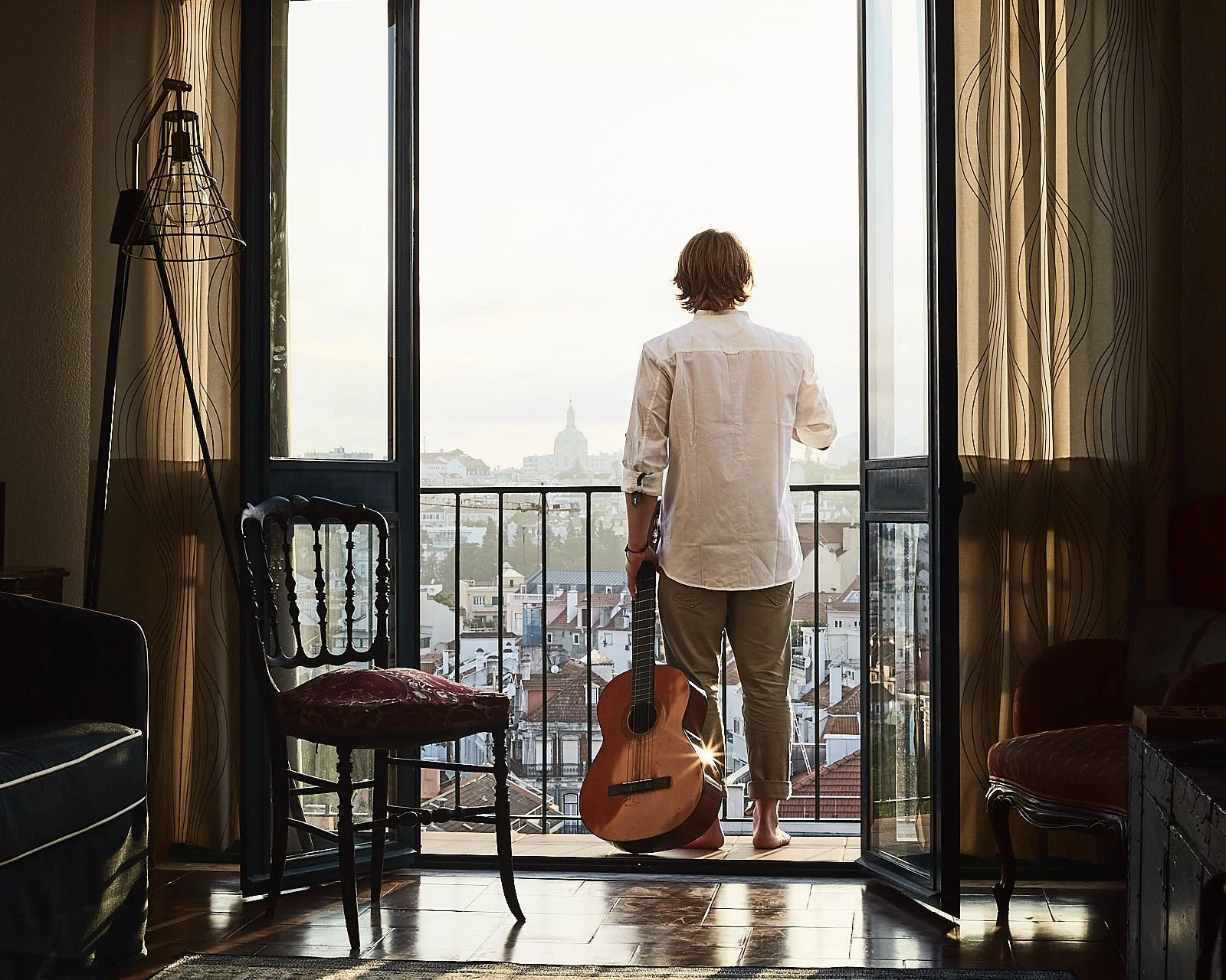 Person standing on a balcony looking at city view, holding a guitar, with a window open and curtains on sides.