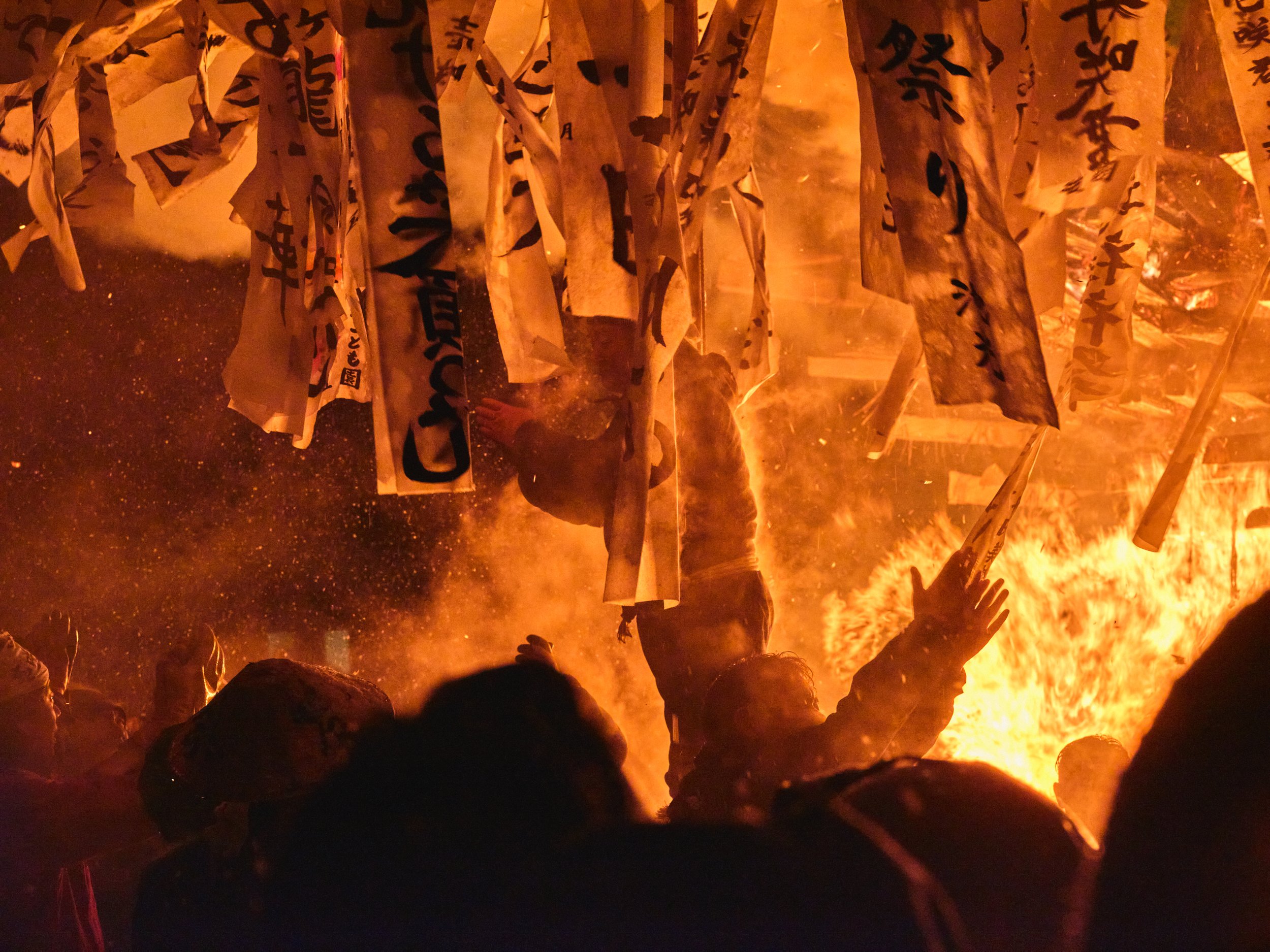 People participating in a traditional Japanese fire festival, surrounded by flames and hanging paper banners with Japanese writing.