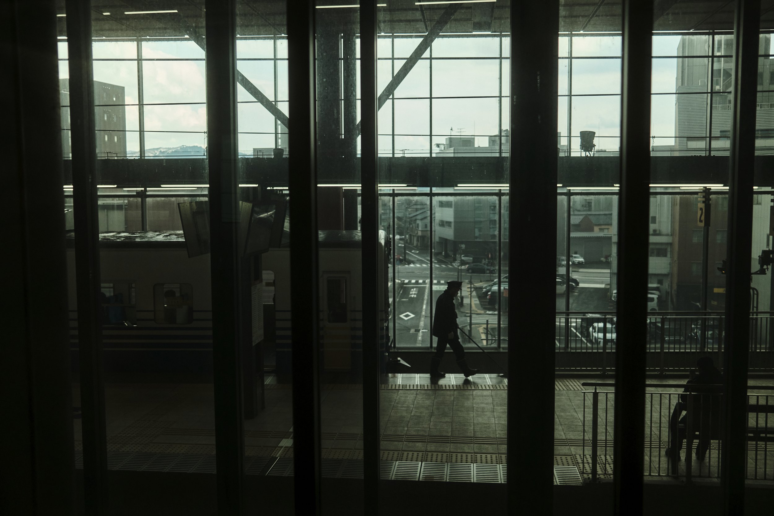 Silhouette of a person walking with a cane inside a modern airport terminal, viewed through tall glass windows showing an urban cityscape with parked cars and buildings.