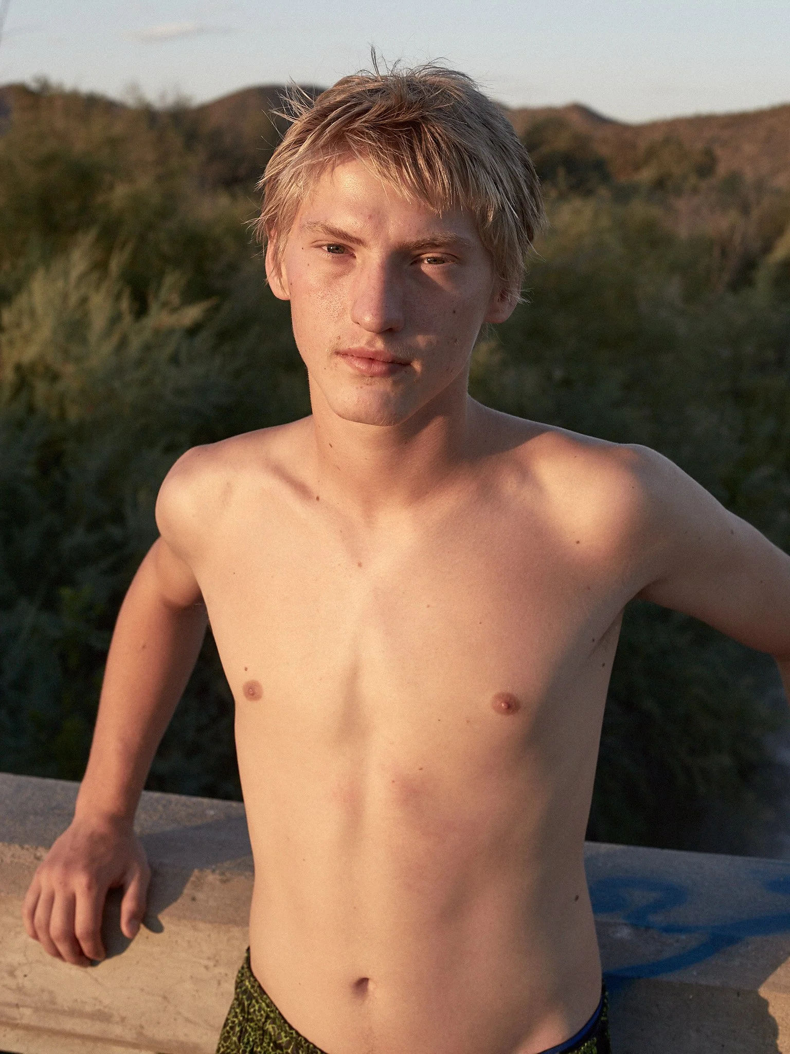 A shirtless young man with light brown hair standing outdoors on a balcony, with mountains and greenery in the background during sunset.