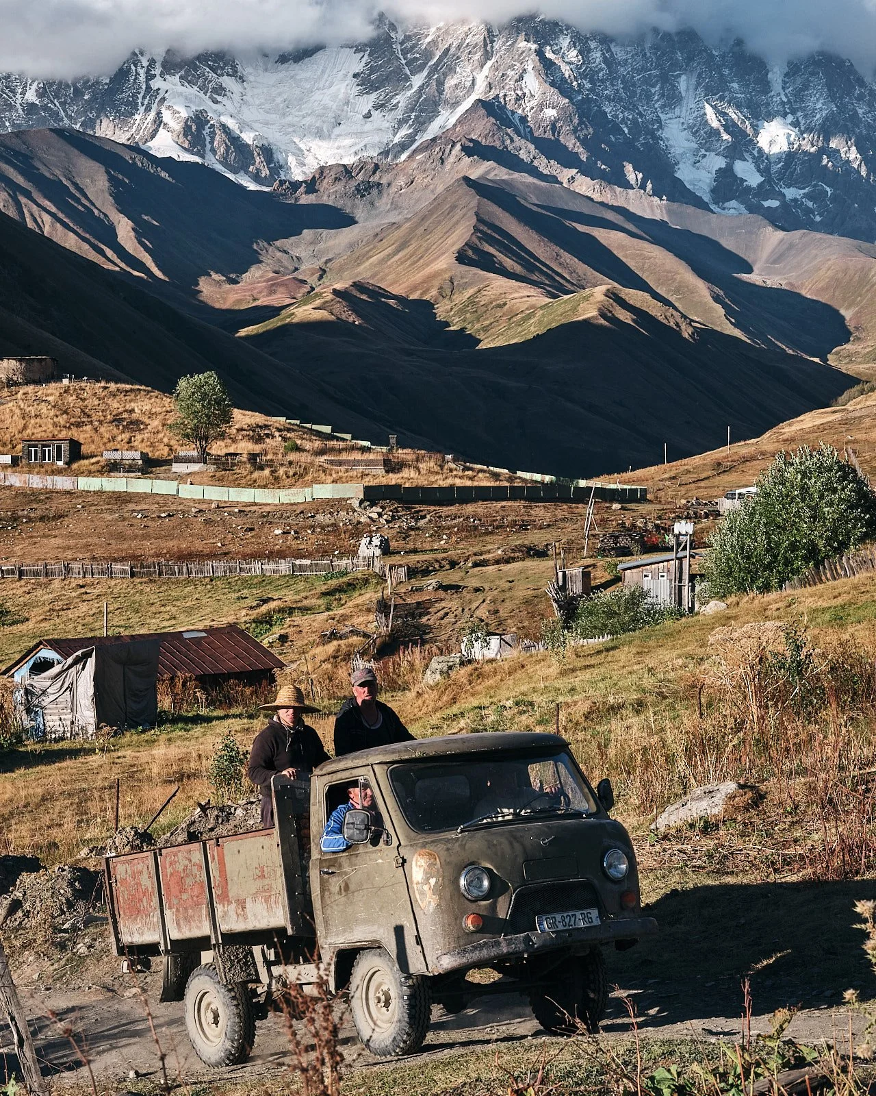 A rugged vintage truck with two people standing in the back, driving through a rural landscape with mountains in the background.