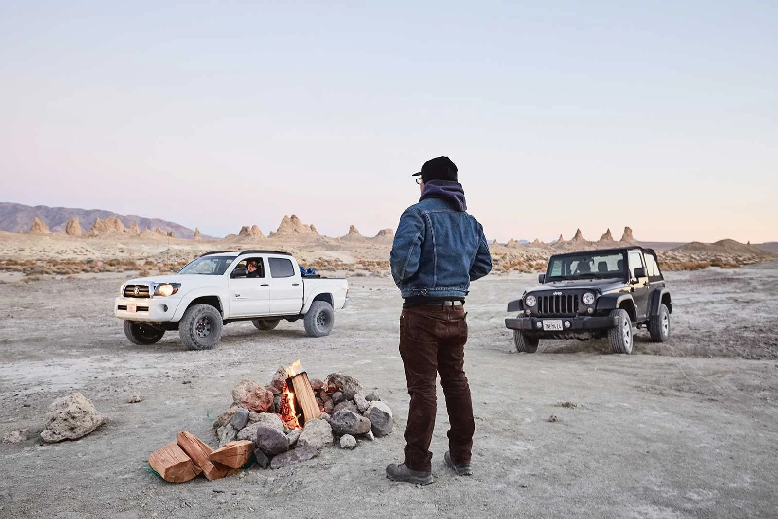 A man standing by a campfire in a desert landscape with two parked off-road vehicles, a white truck and a black Jeep, during sunset or sunrise.