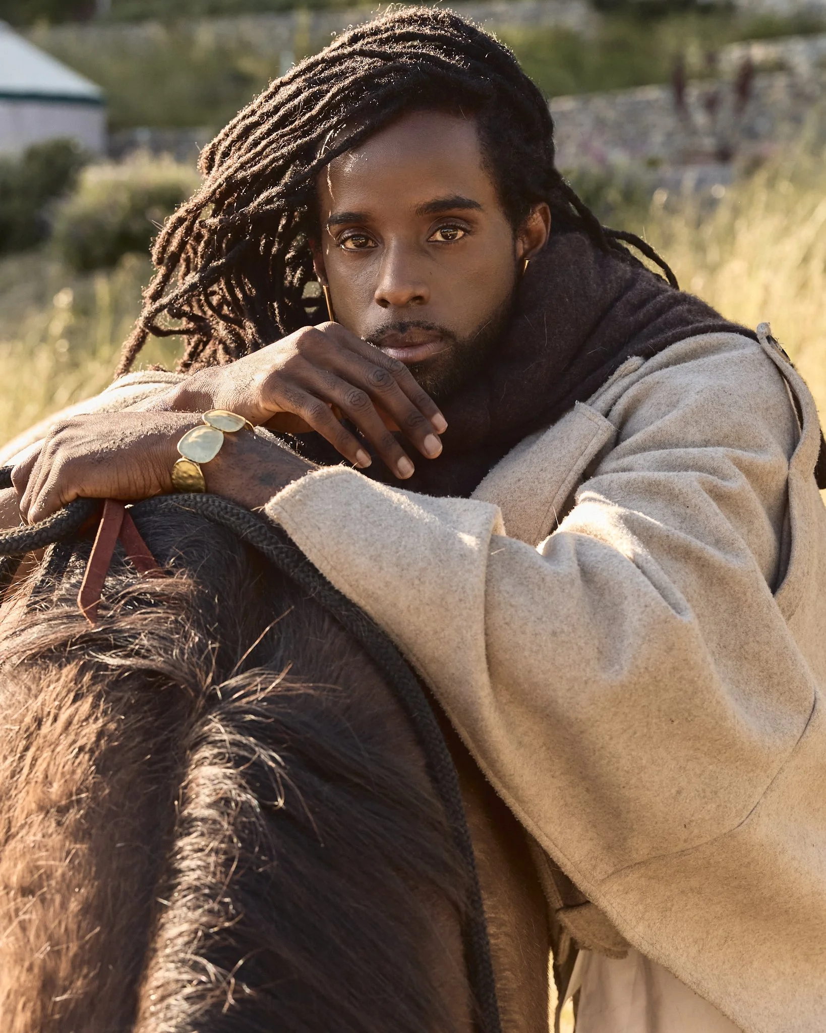 A man with dreadlocks is resting his head and arm on a horse's neck, looking into the camera with a serious expression, outdoors during the daytime.