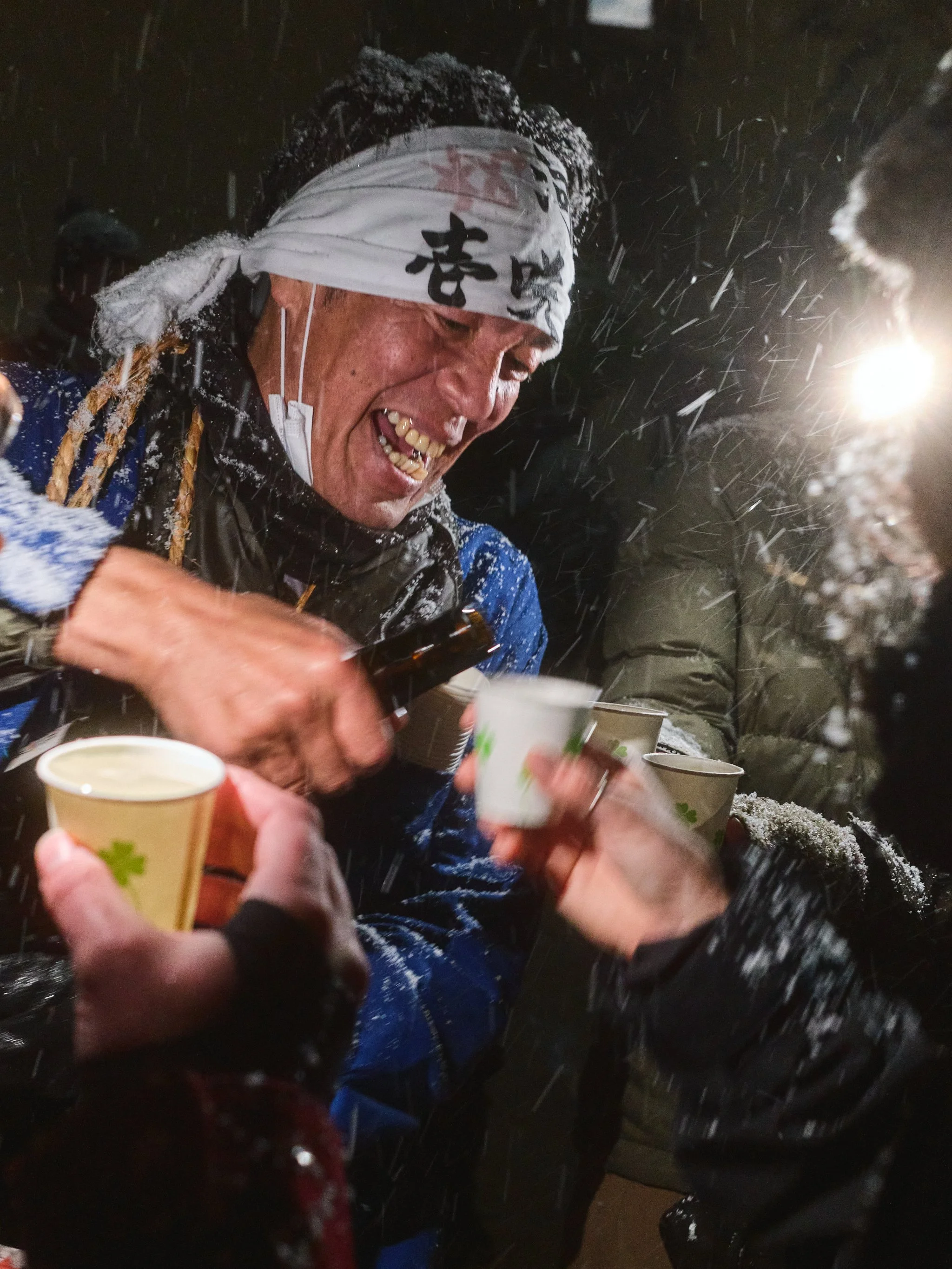 A joyful man wearing a headband with Japanese characters, laughing and serving drinks to others in a snowy outdoor setting at night.