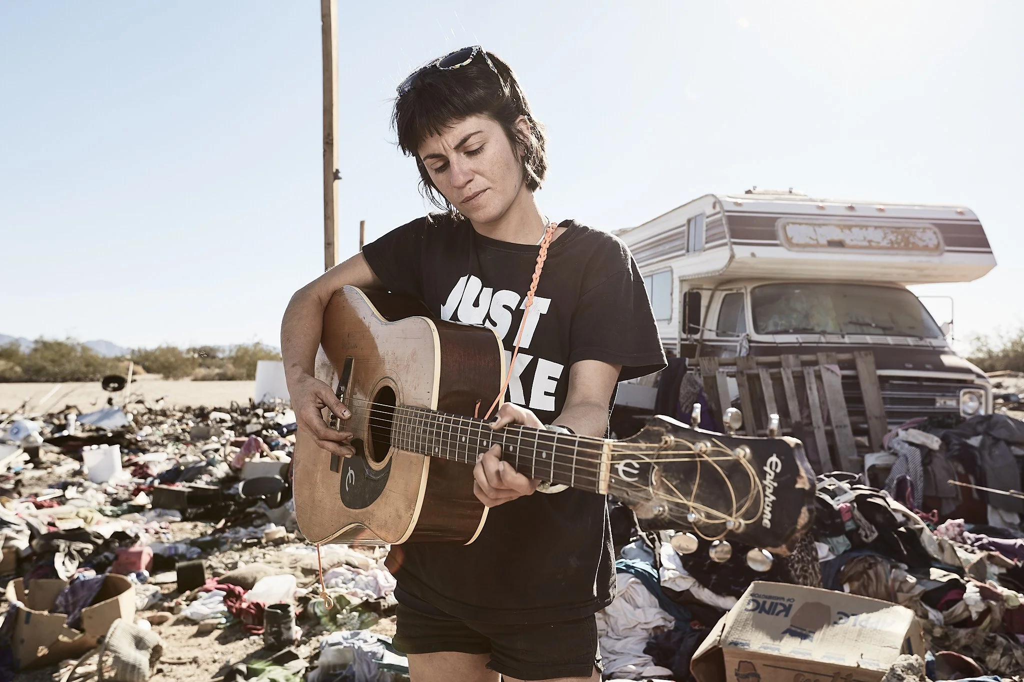A young woman with short dark hair playing an acoustic guitar outdoors amid a large pile of trash, with a motorhome in the background.