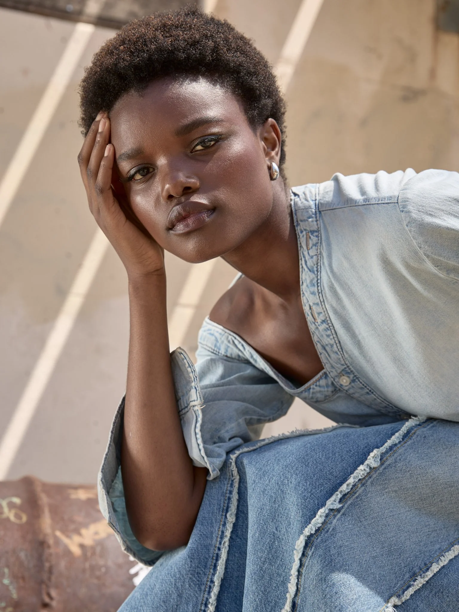 A young woman with short curly hair, wearing a light denim off-shoulder top and distressed jeans, sitting outdoors with an intense expression, resting her head on her left hand.