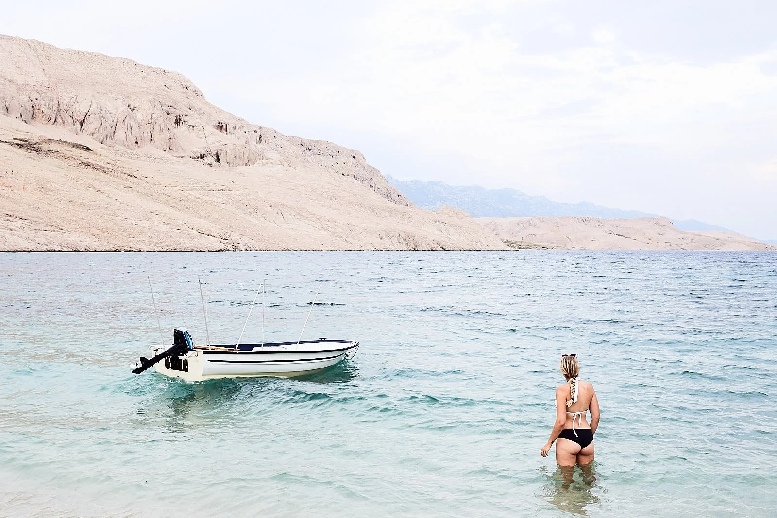 A woman with a swimsuit and a towel around her waist wading in the water near a small boat on a calm lake with mountains in the background.