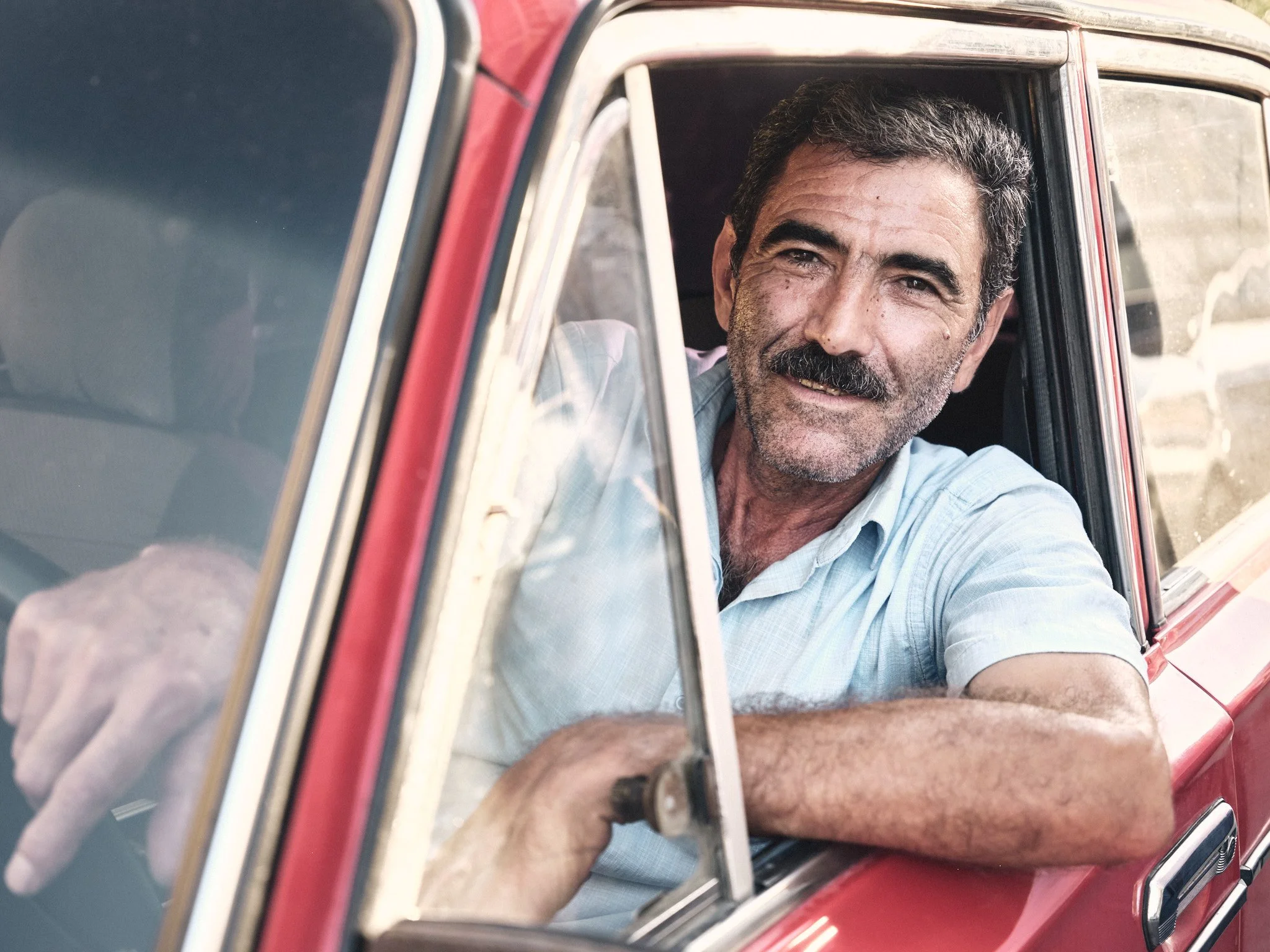 A middle-aged man with a beard and mustache, smiling, sitting in the driver's seat of a red car, resting his arm on the window ledge, wearing a light blue shirt.