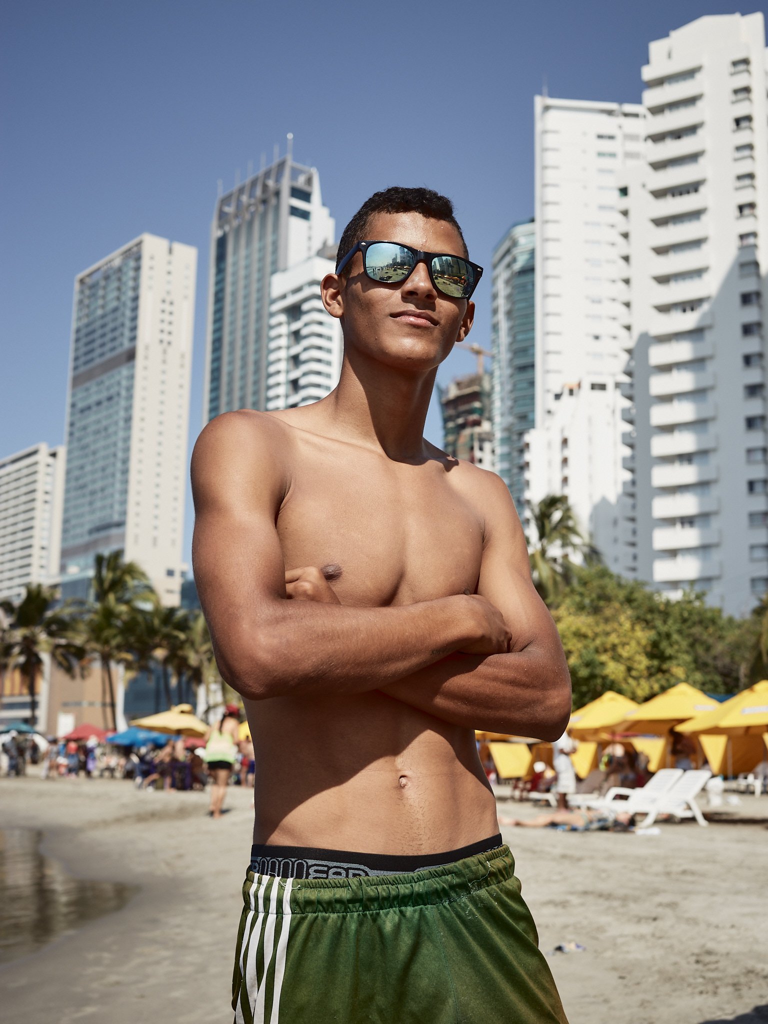 A shirtless young man wearing sunglasses and green striped shorts standing with arms crossed on a busy beach with tall buildings and yellow umbrellas in the background.