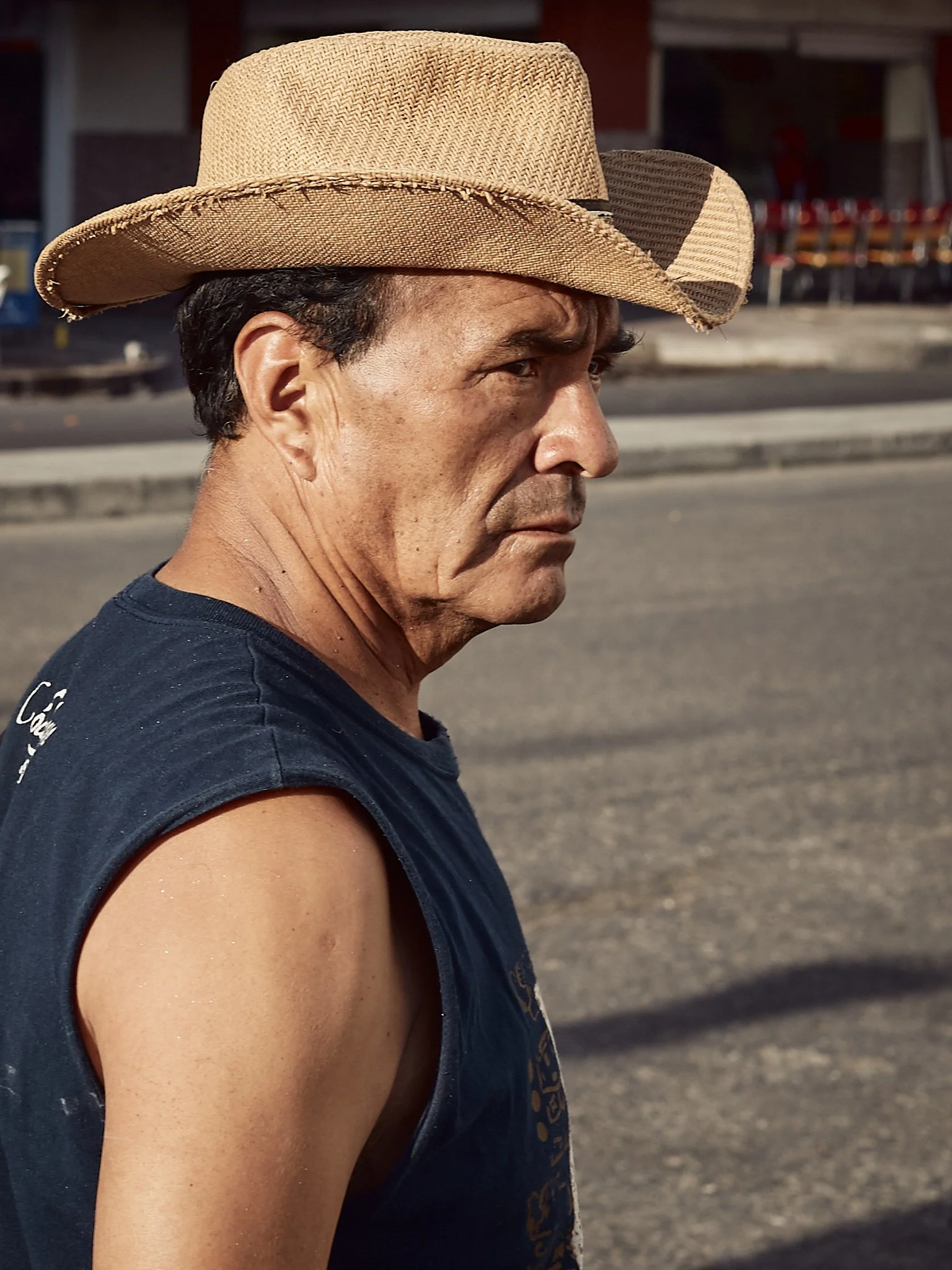 Side profile of a man wearing a sleeveless dark T-shirt and a straw cowboy hat, standing outdoors with a blurred background of a road and storefronts.