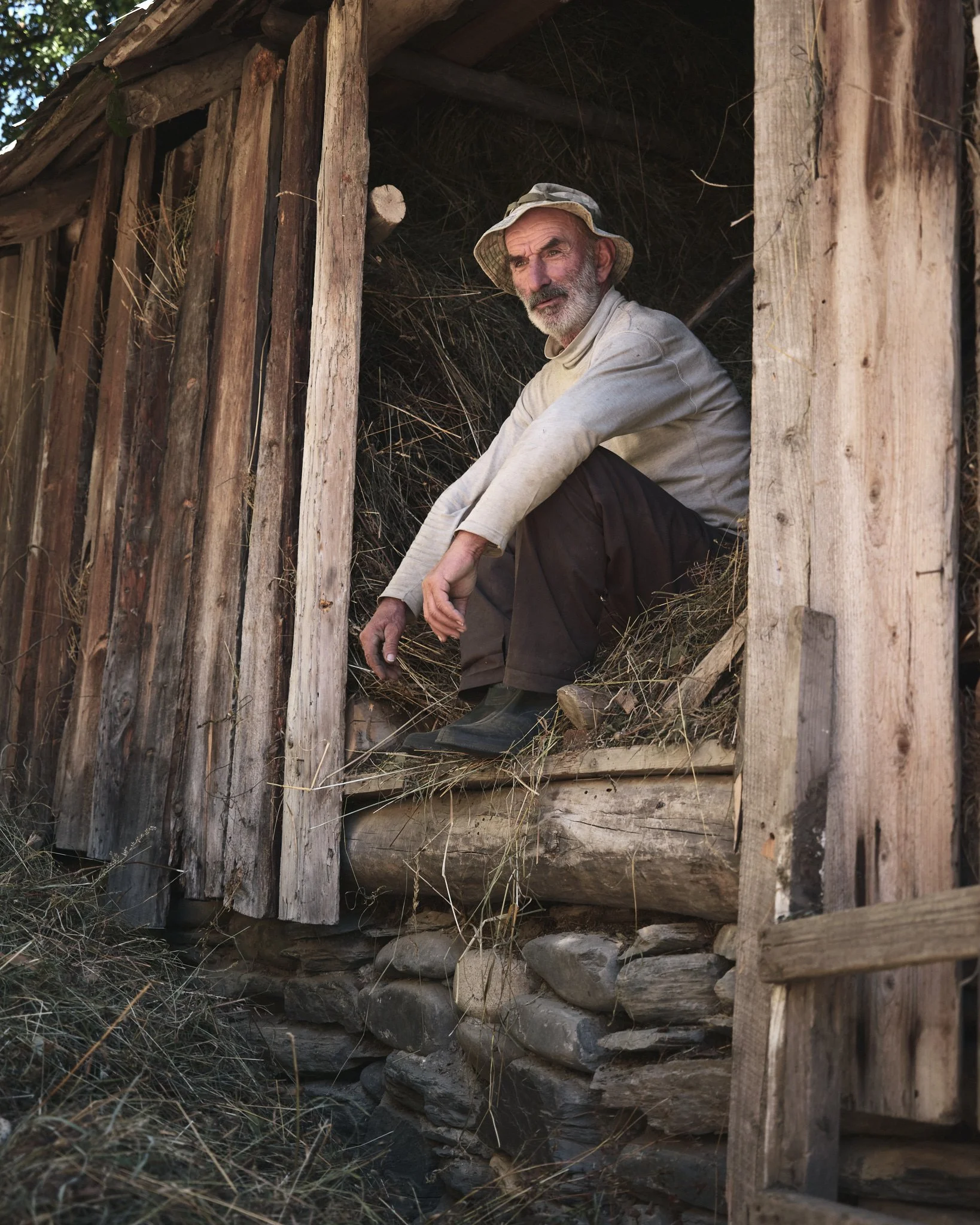 An elderly man sitting inside a rustic wooden shelter with a stone foundation, wearing a hat, beige shirt, and dark pants, looking pensively outside.