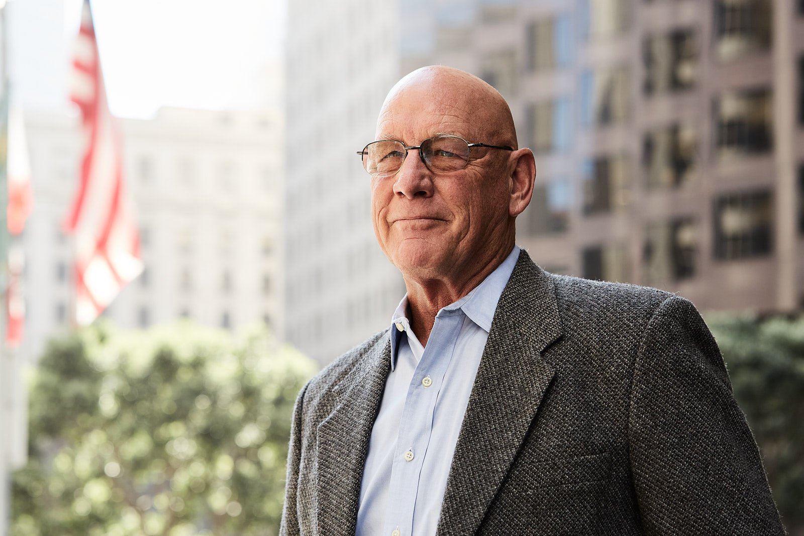 A middle-aged man in glasses and a gray blazer standing outdoors in front of a cityscape with tall buildings and American flags.