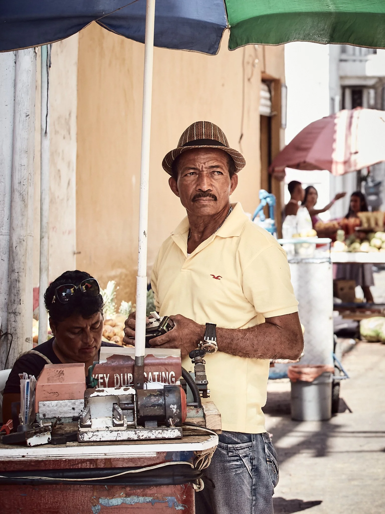 A man wearing a striped hat and yellow shirt stands next to a street vendor table, holding a camera, with a woman seated behind the table under a colorful umbrella at an outdoor market.