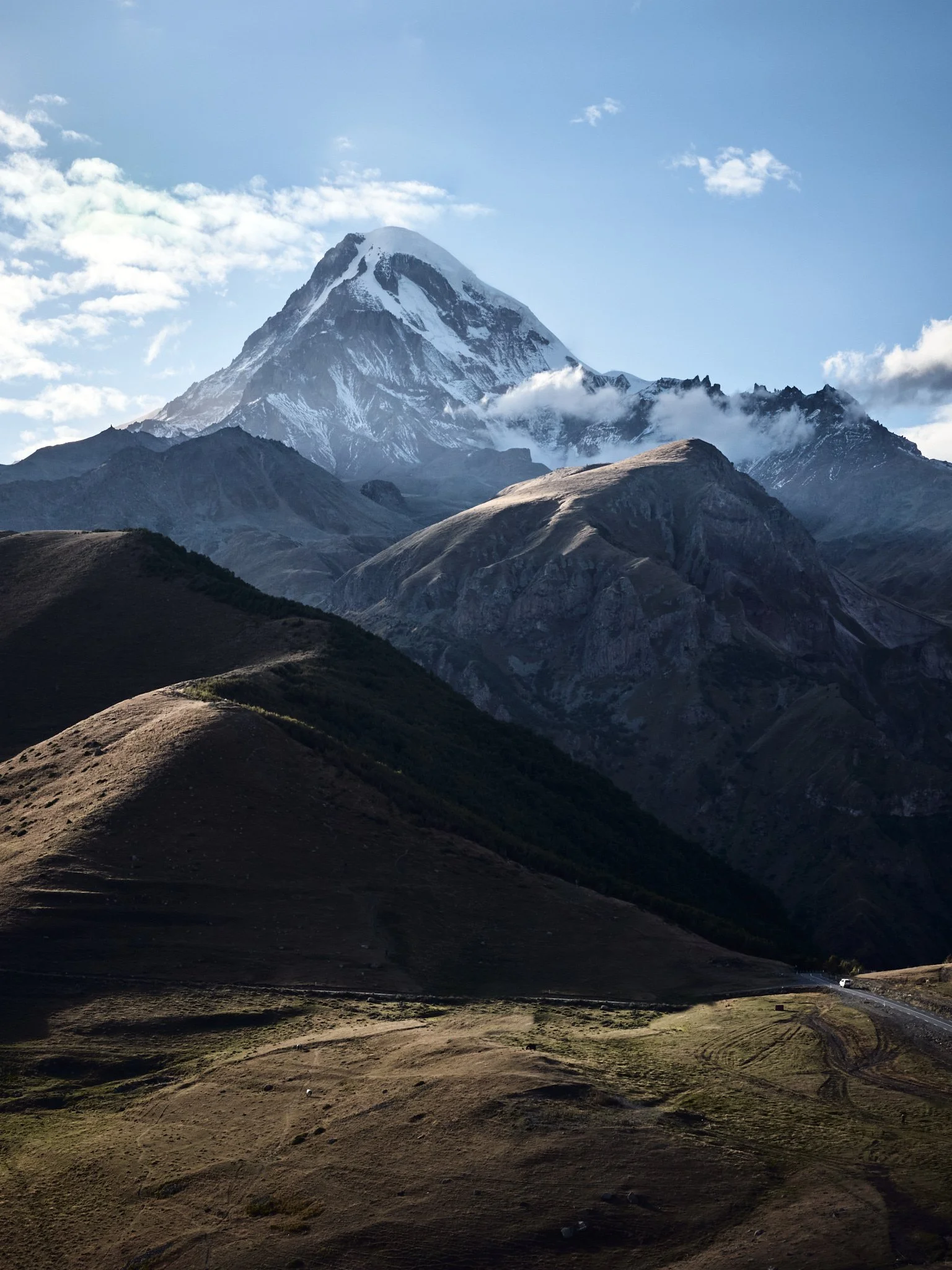 Snow-capped mountain peak soaring above rolling grassy hills and rugged terrain under a partly cloudy sky.