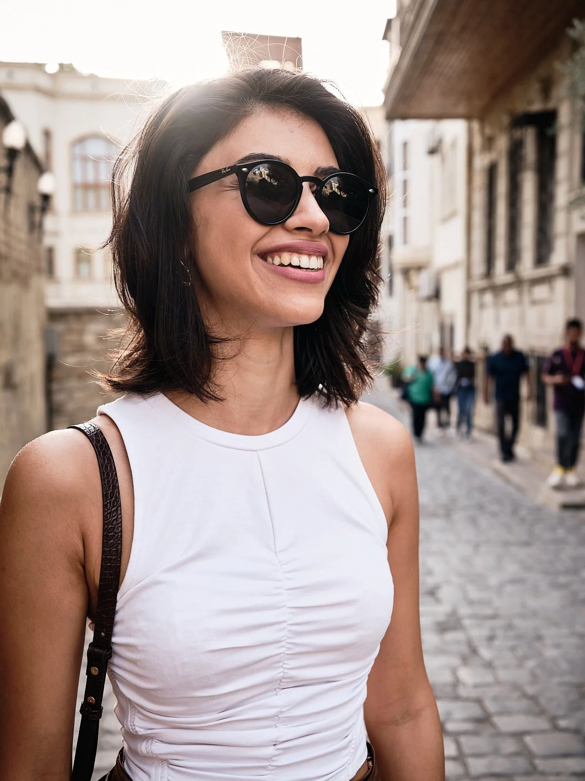 A smiling woman with shoulder-length dark hair wearing sunglasses and a white sleeveless top, standing on a cobblestone street in an urban area.