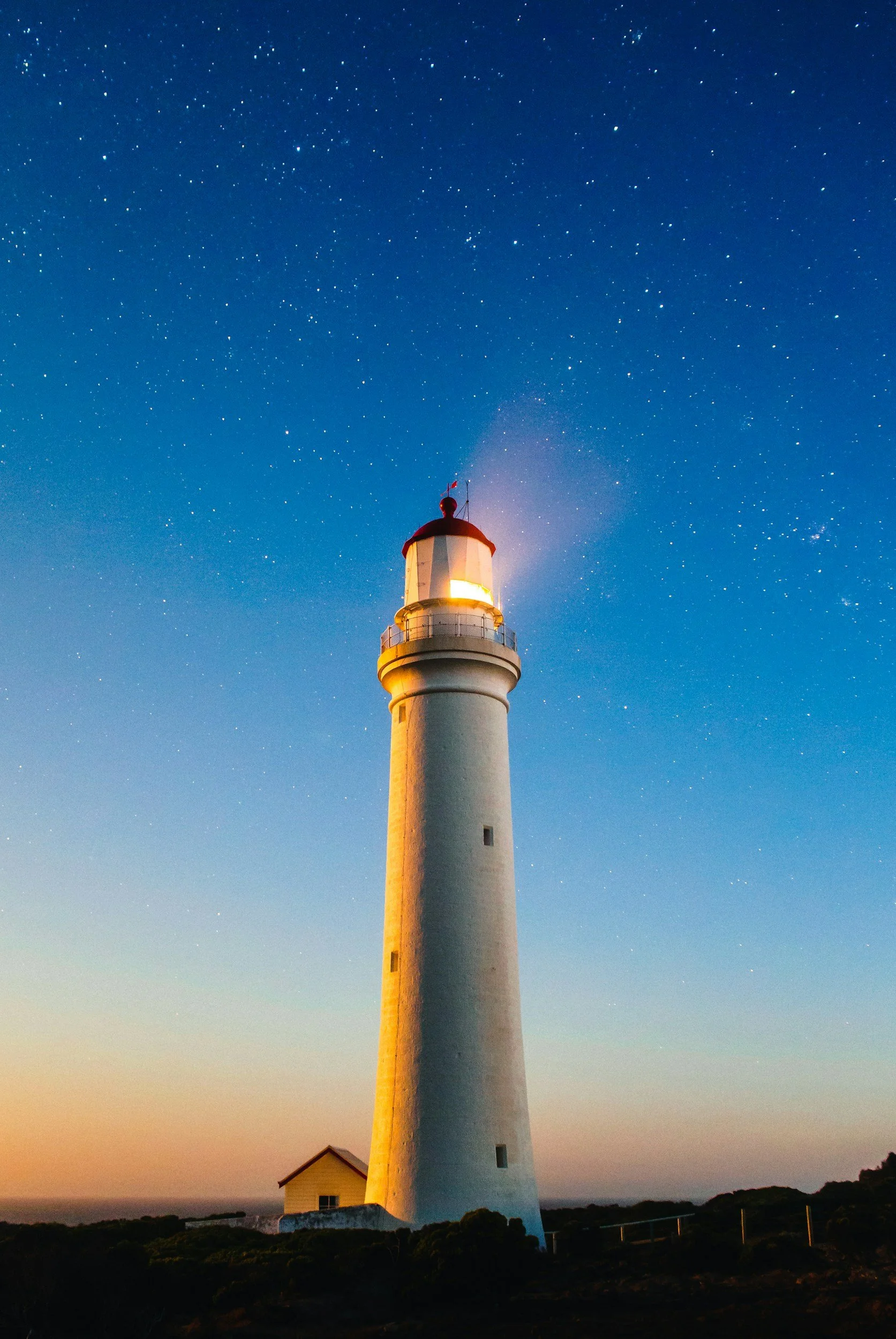 A tall white lighthouse with a red roof and balcony, illuminated at night against a starry sky.