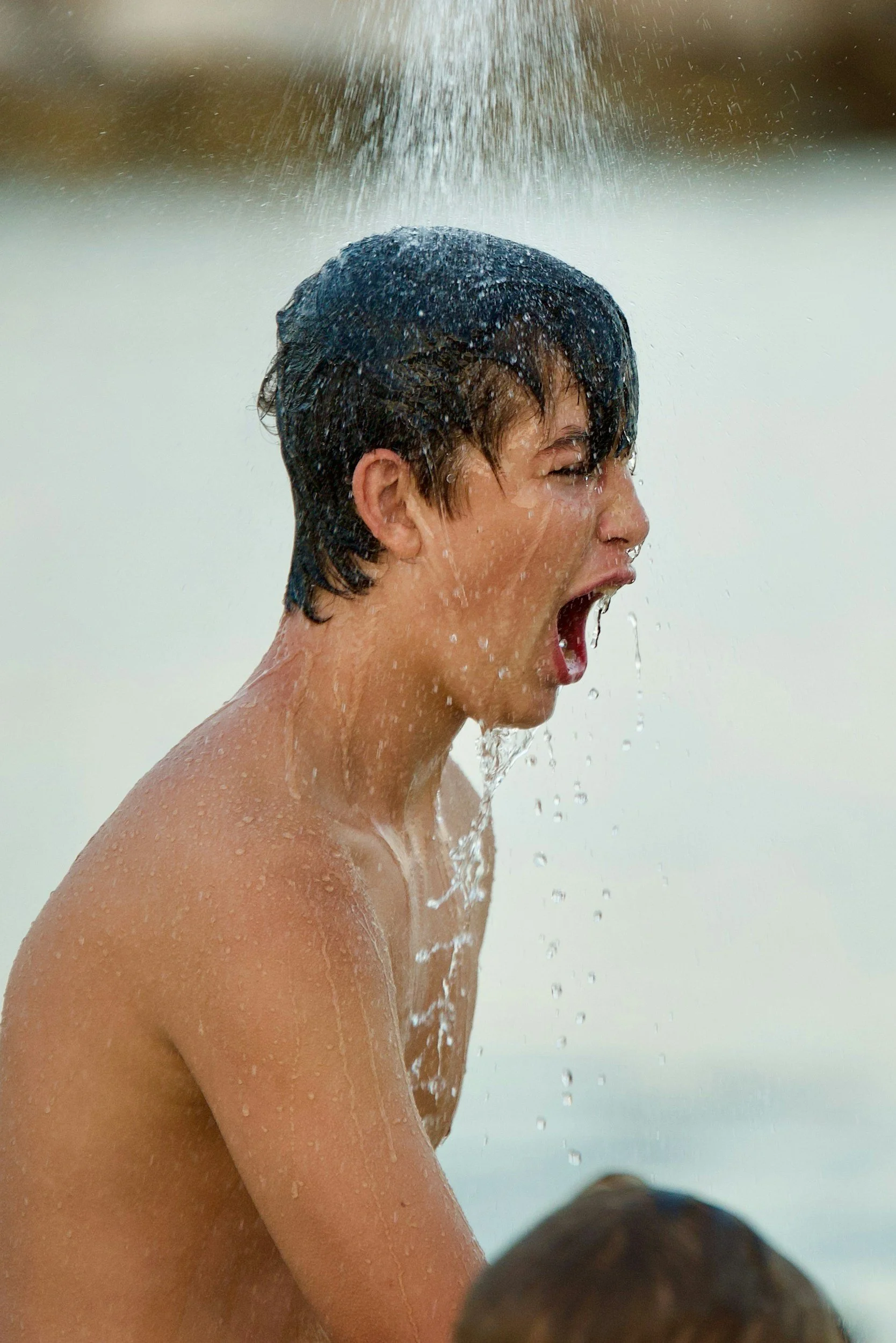 A young boy with wet hair, water splashing on his face, mouth open, appearing to shout or yell, outdoors near water.