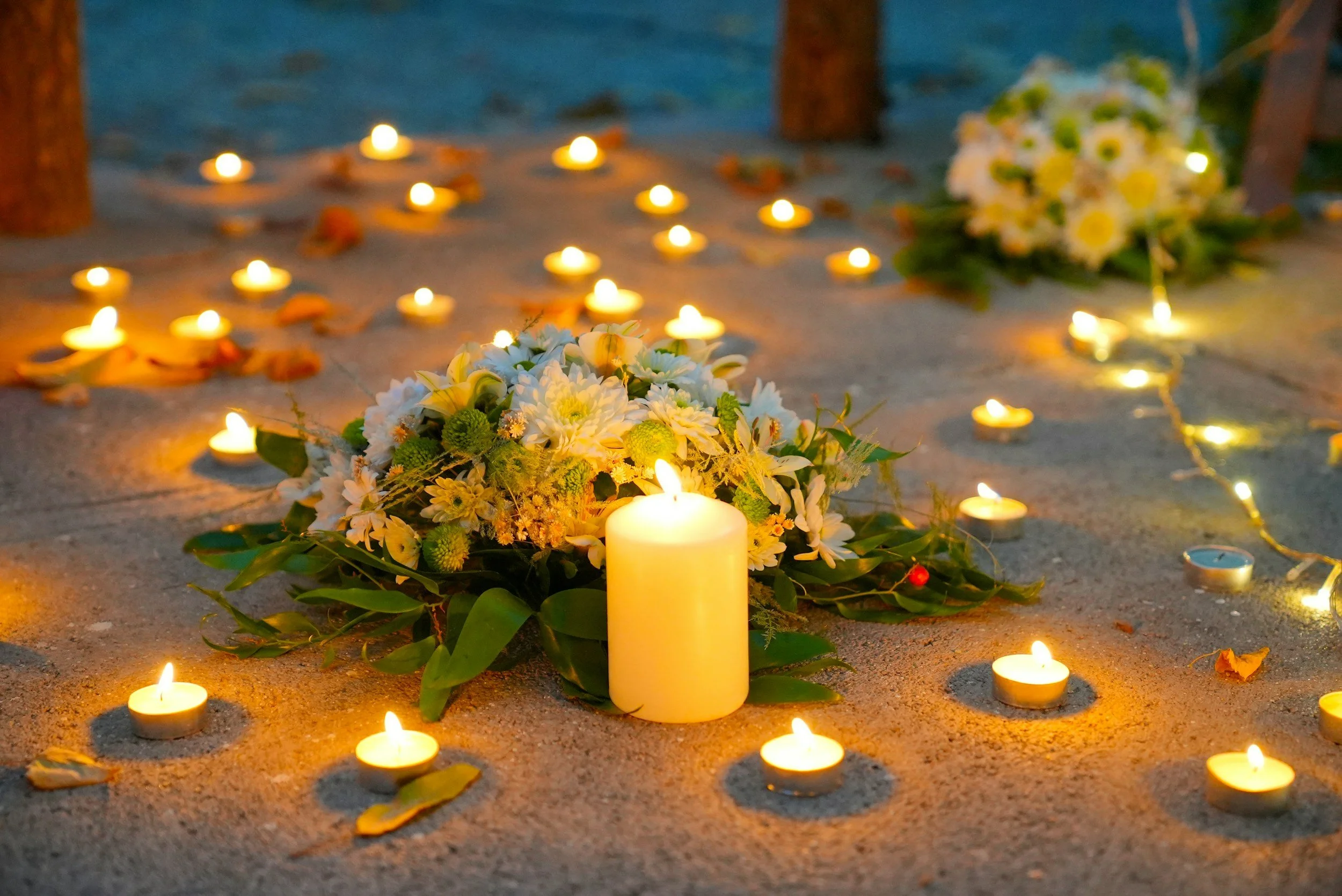 An evening memorial setup with candles, flowers, and leaf arrangements on the ground.