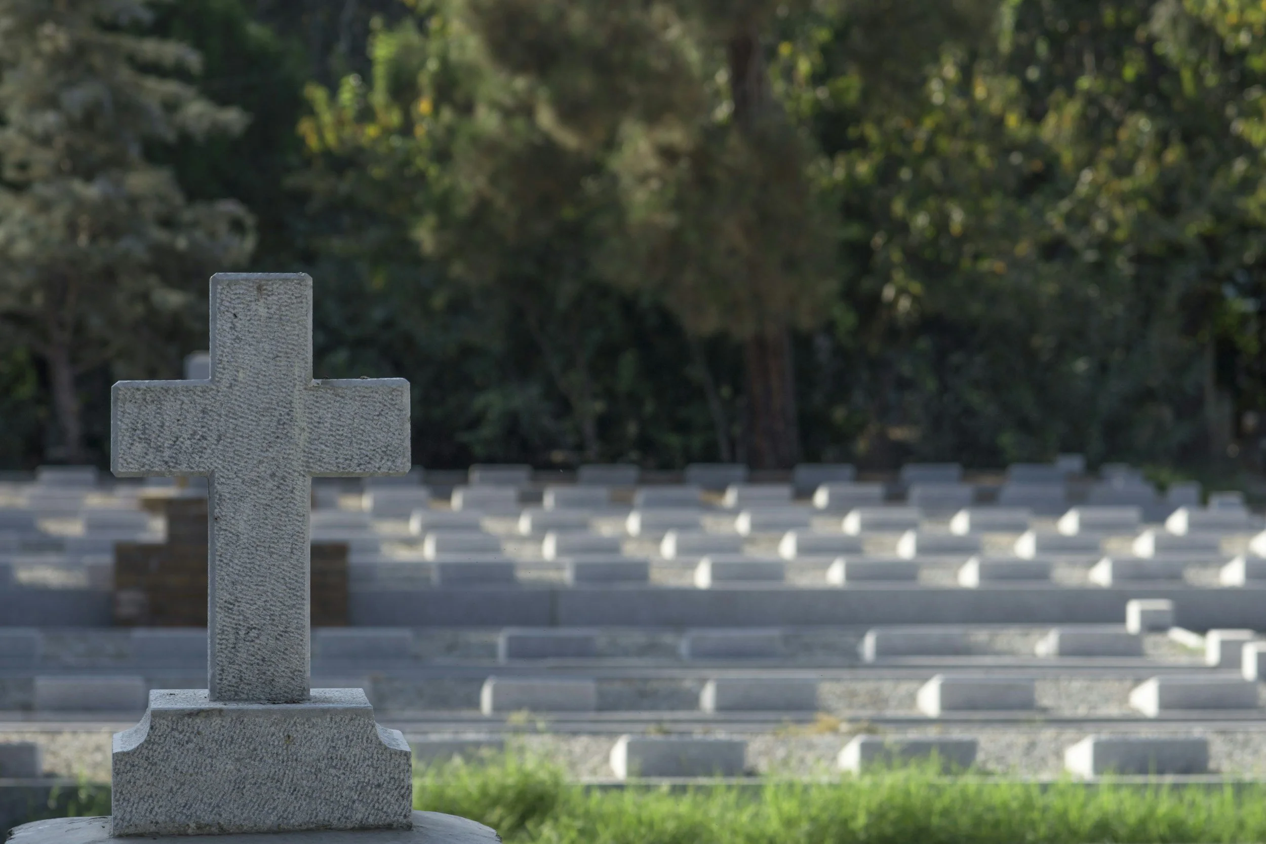 A gray stone cross monument with engraved text in the foreground, with rows of similar stone crosses in the background in a cemetery, surrounded by trees.