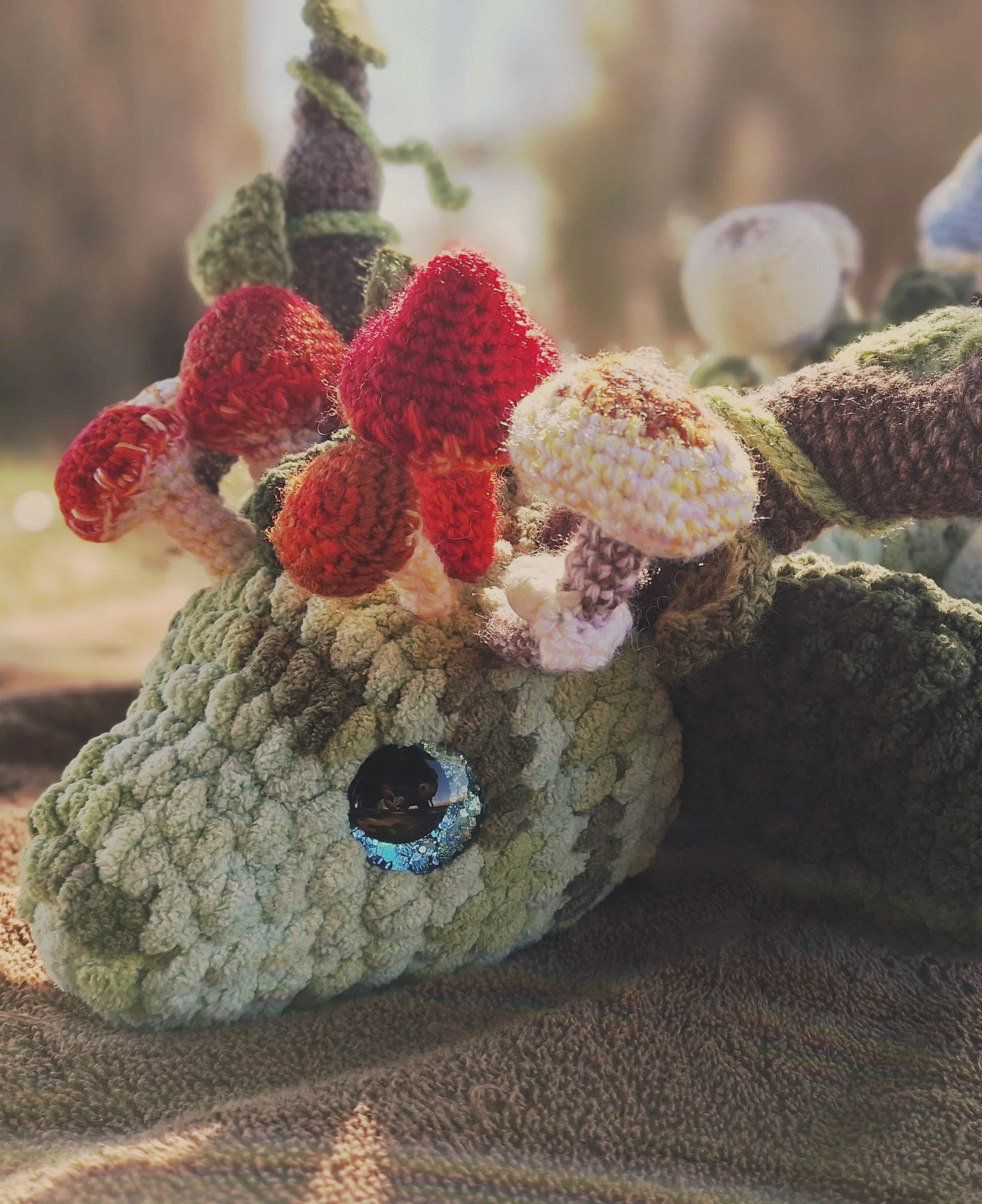 A crochet, wingless forest dragon with gentle blue eyes that shimmer like the deepest rain forest pools and mushrooms growing around her horns and across her whole body. Mossy stones and leaves can also be seen on her legs and tail.