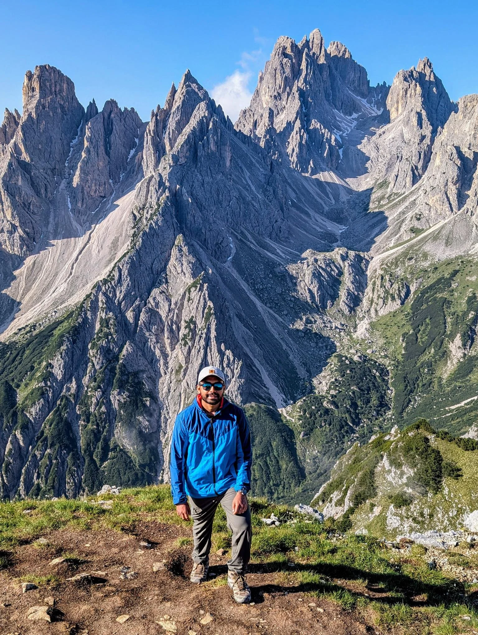 A man wearing sunglasses, a blue outdoor jacket, and a baseball cap standing on a grassy trail with towering rocky mountains in the background under a clear blue sky.