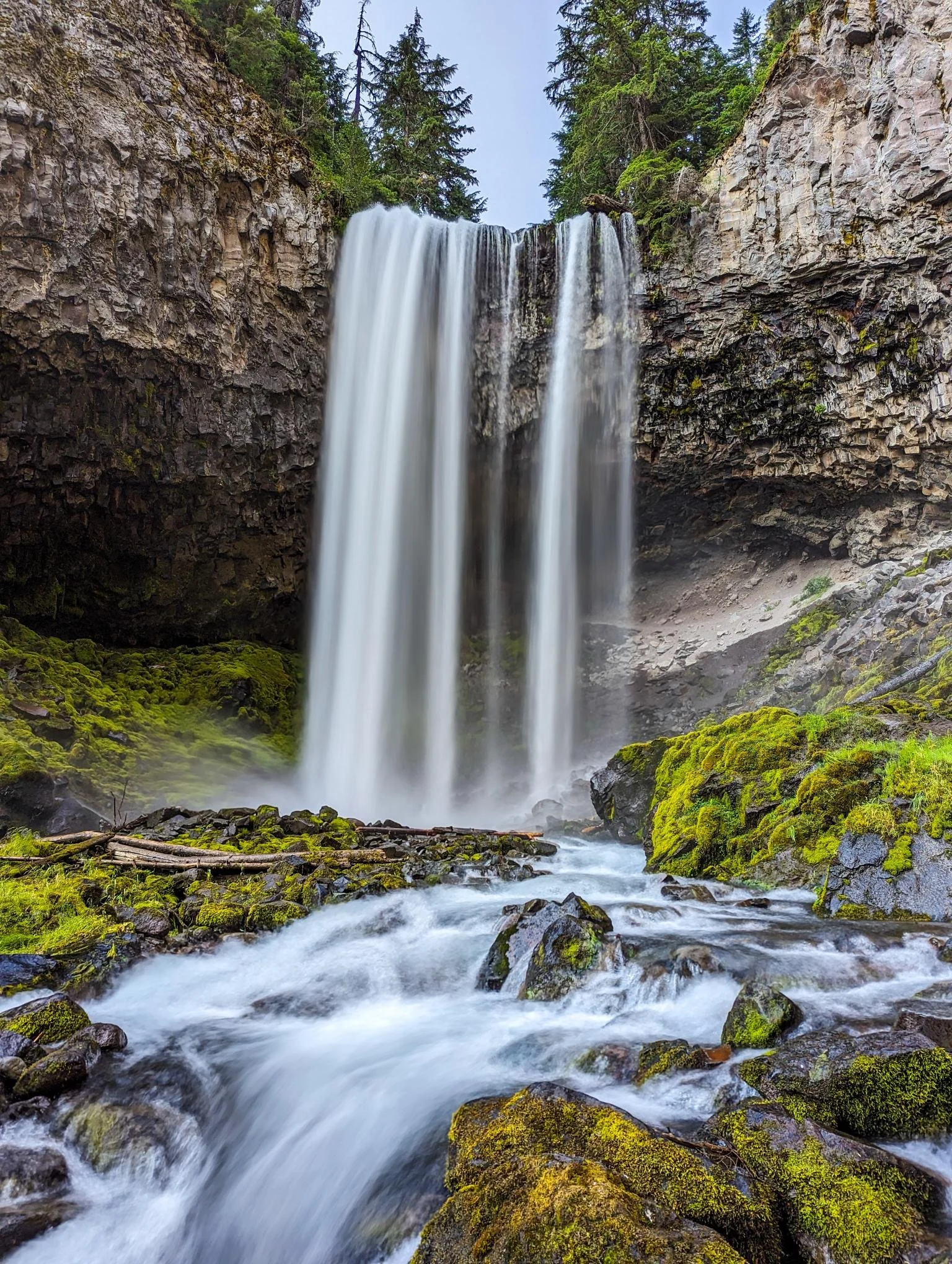 A tall waterfall flowing over a cliff surrounded by dense green trees, with water cascading into a moss-covered rocky stream below.