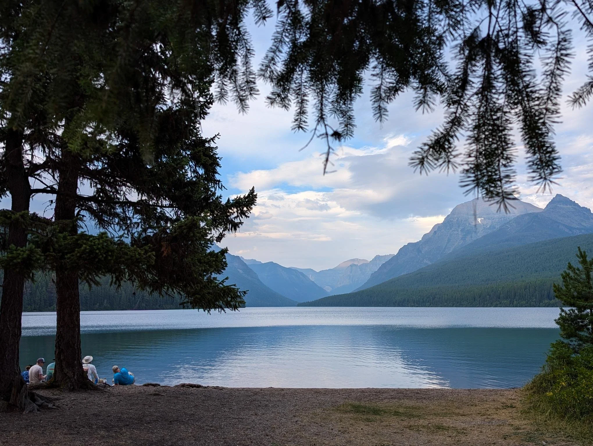 A group of people sitting on the shore of a lake surrounded by tall trees and mountains in the background, with partly cloudy sky.