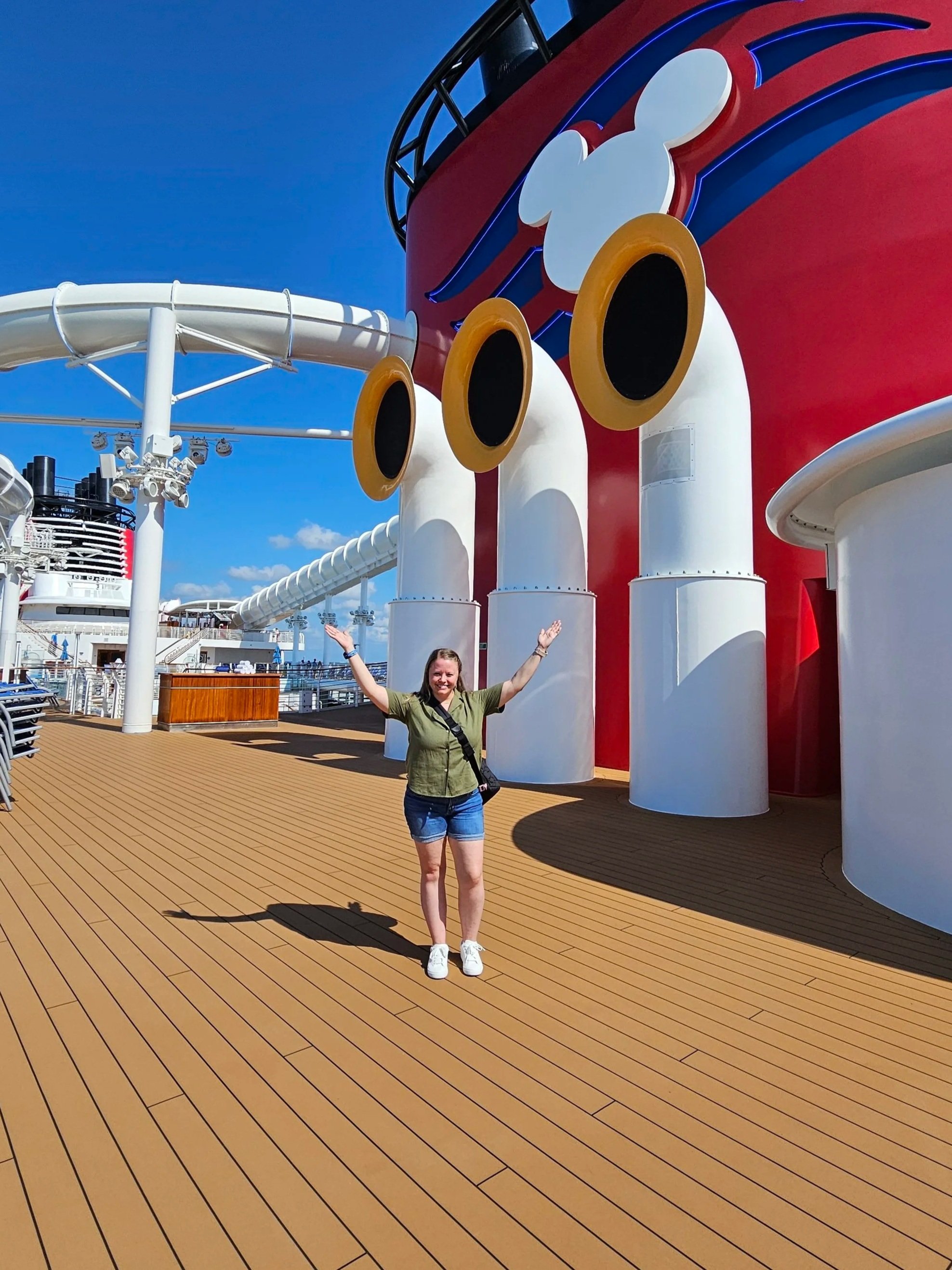 A woman standing on the deck of a cruise ship with her arms raised, in front of a red and white themed area with large portholes and a Mickey Mouse silhouette.