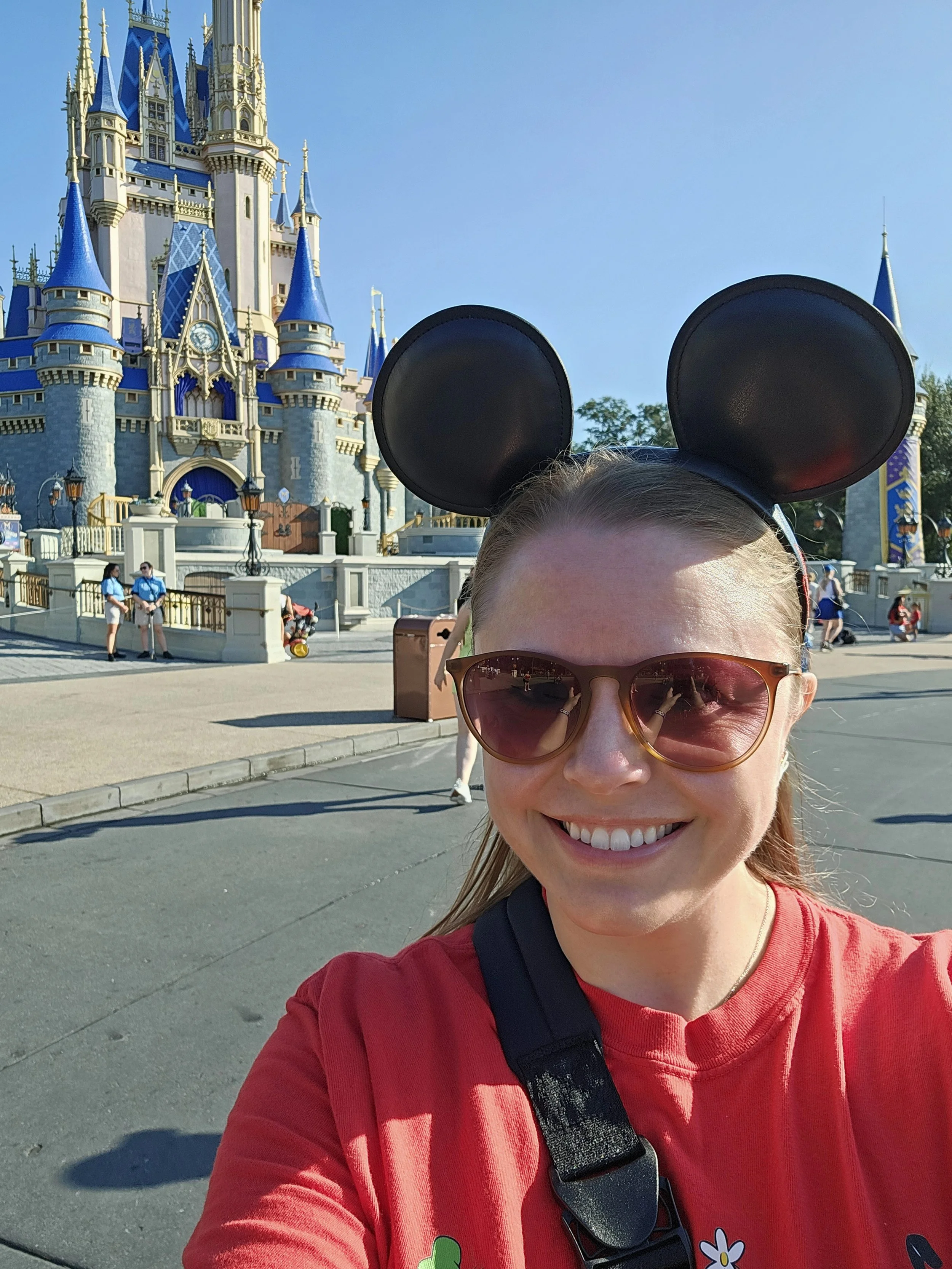 A woman smiling in front of Sleeping Beauty Castle at Disneyland, wearing Mickey Mouse ears and sunglasses.
