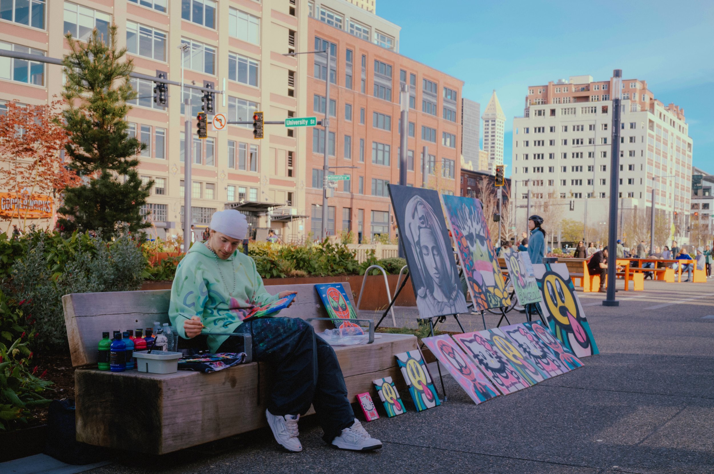 An artist sitting on a park bench by his colorful paintings, with buildings and street signs in the background.