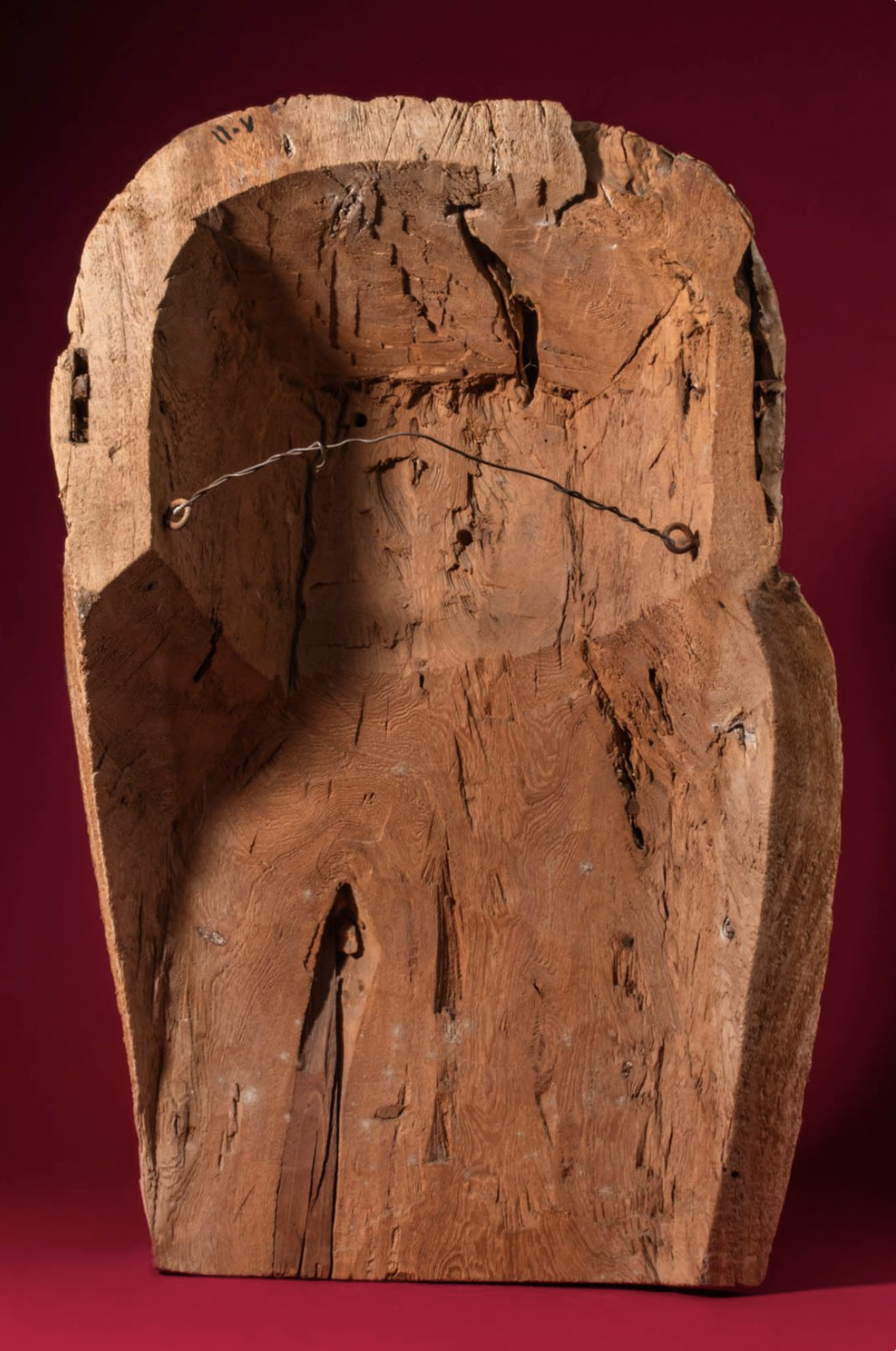 A wooden carved statue viewed from the back, with visible wood grain, cracks, and a metal wire fastener.